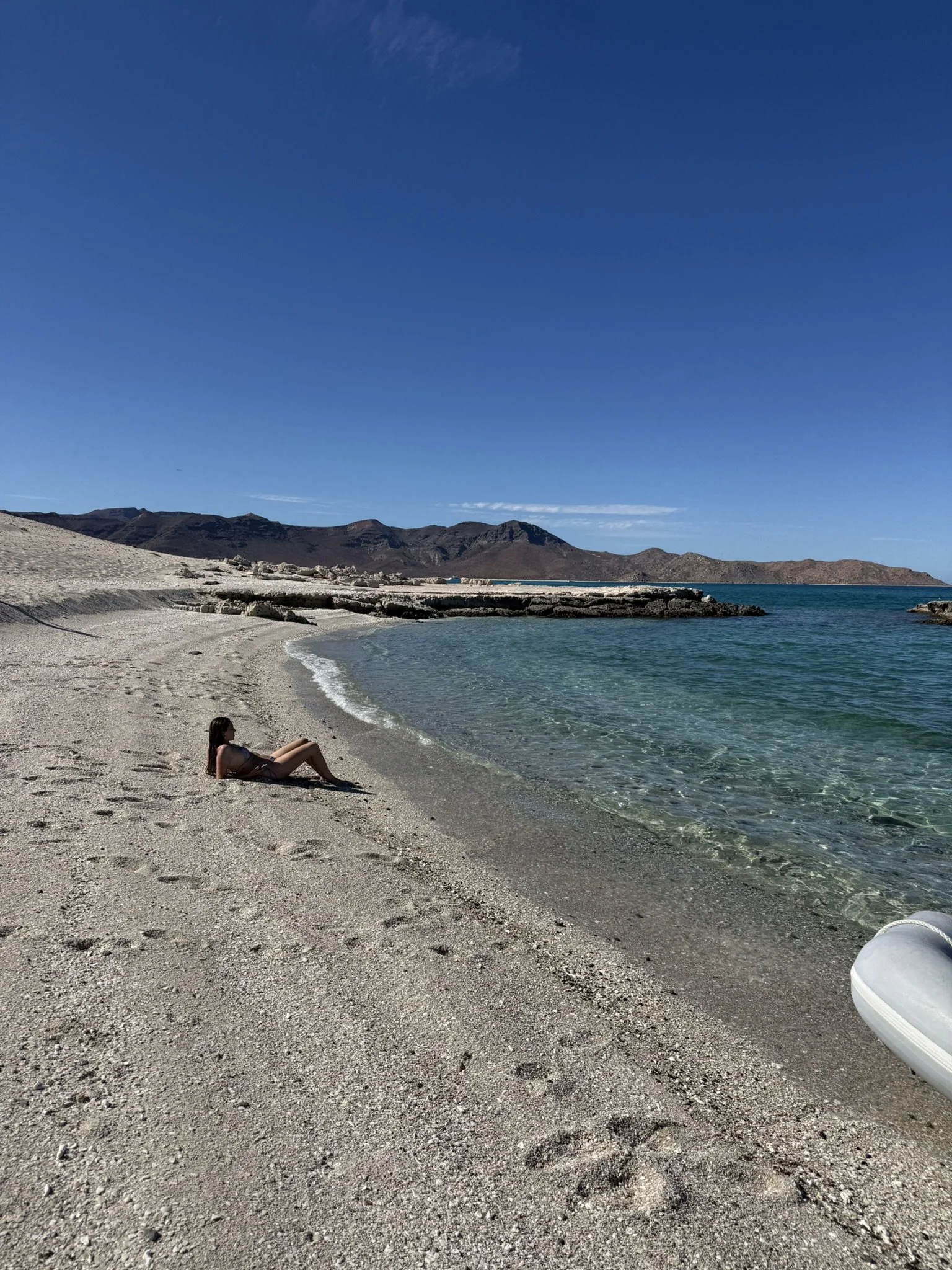 A woman relaxing on a sandy beach with clear blue water and mountains in the background under a blue sky with a few clouds.