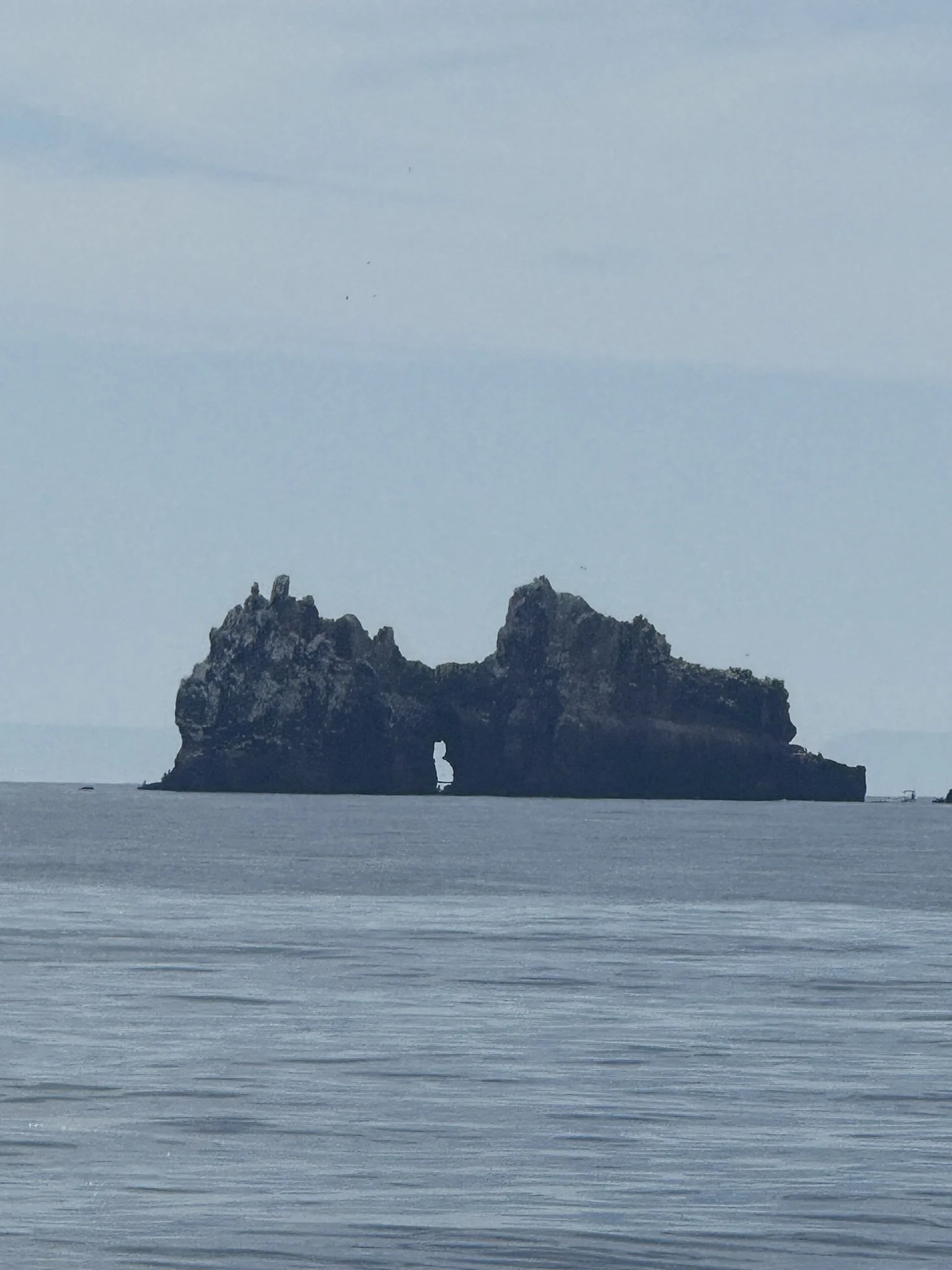A large rock formation in the water with a natural arch, under a cloudy sky, in a coastal scene.