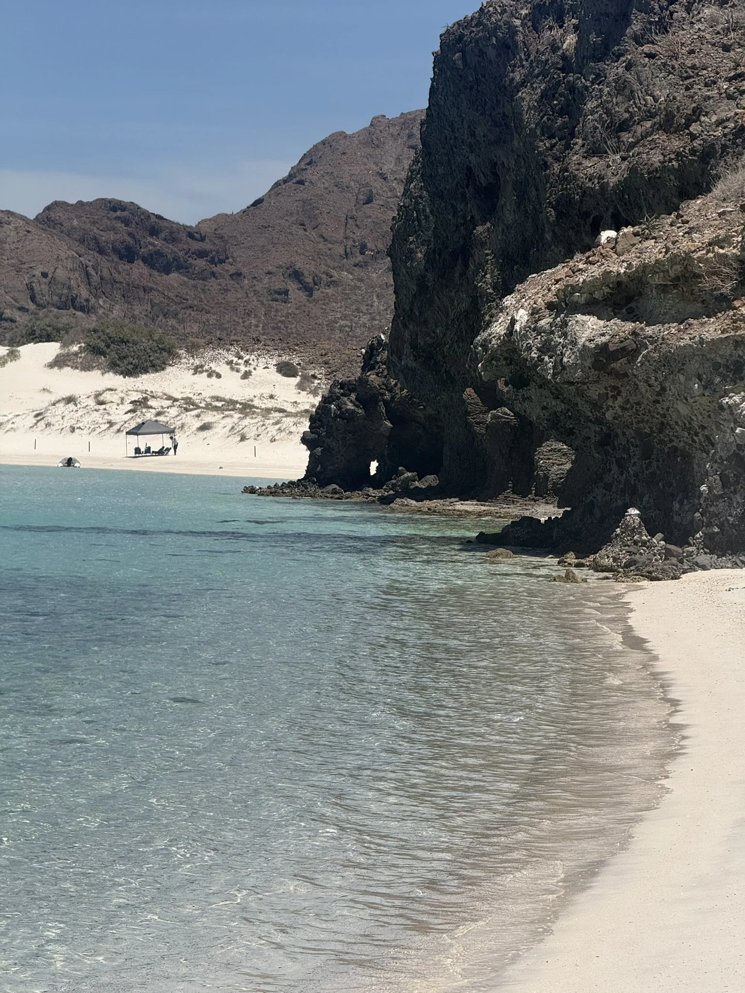 A secluded beach with white sand, turquoise water, rocky cliffs, and mountains in the background, with a shaded hammock and chairs on the sand.