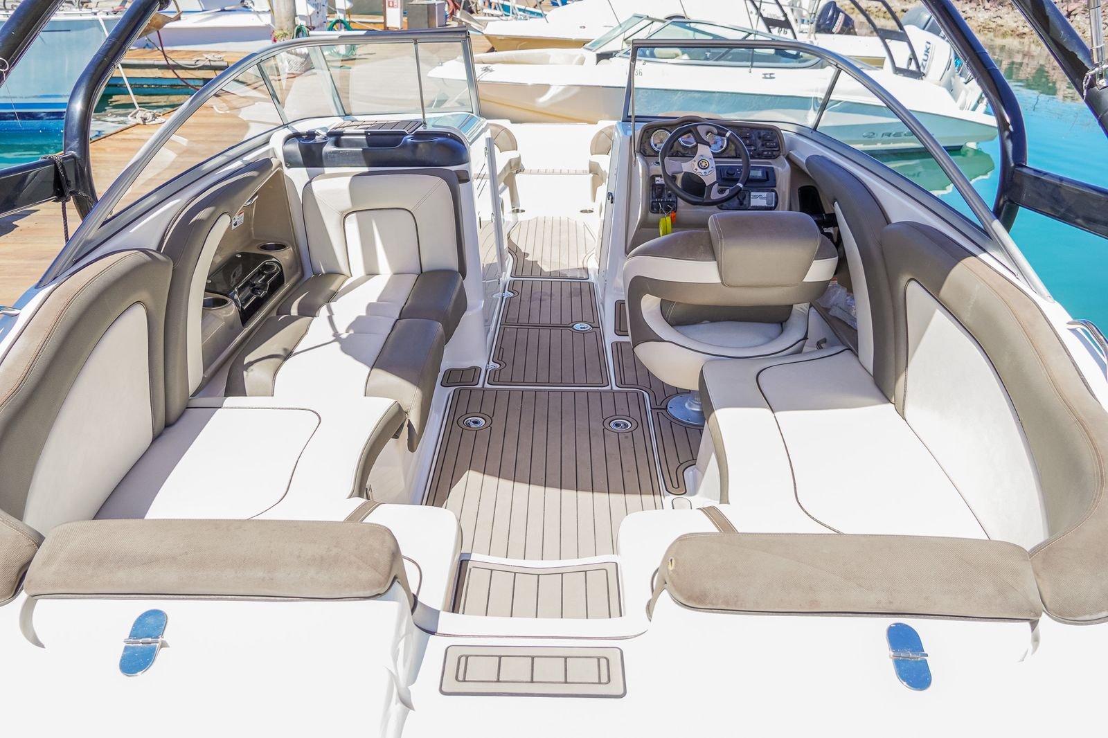 Interior of a boat with beige cushioned seats, a steering wheel, and a dashboard, docked at a marina with other boats and calm water visible.