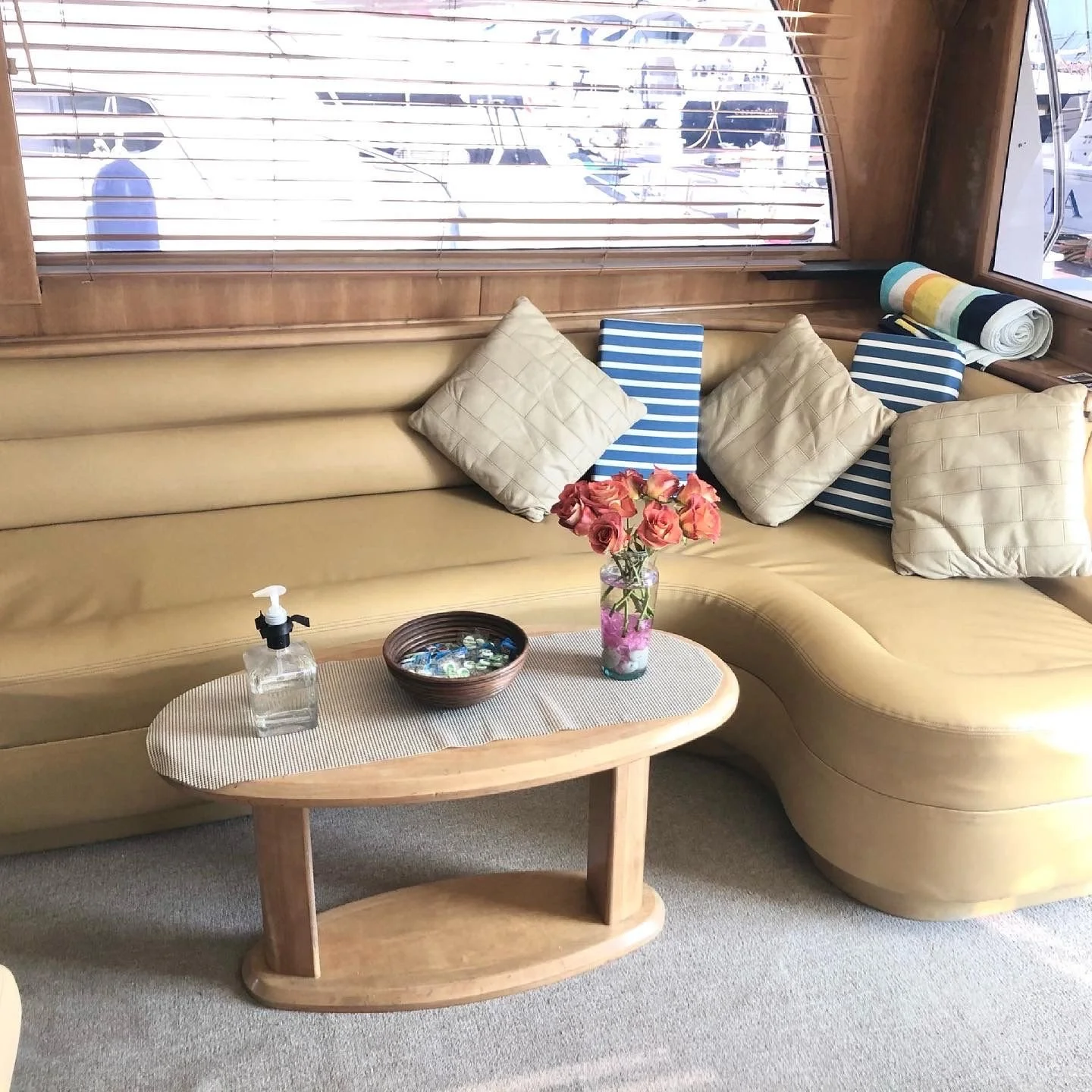 A cozy seating area with a curved beige sofa decorated with several beige and striped pillows, a wooden coffee table with a vase of pink roses, a bowl of shells, and a hand sanitizer dispenser. Behind the sofa, there are windows with wooden blinds re