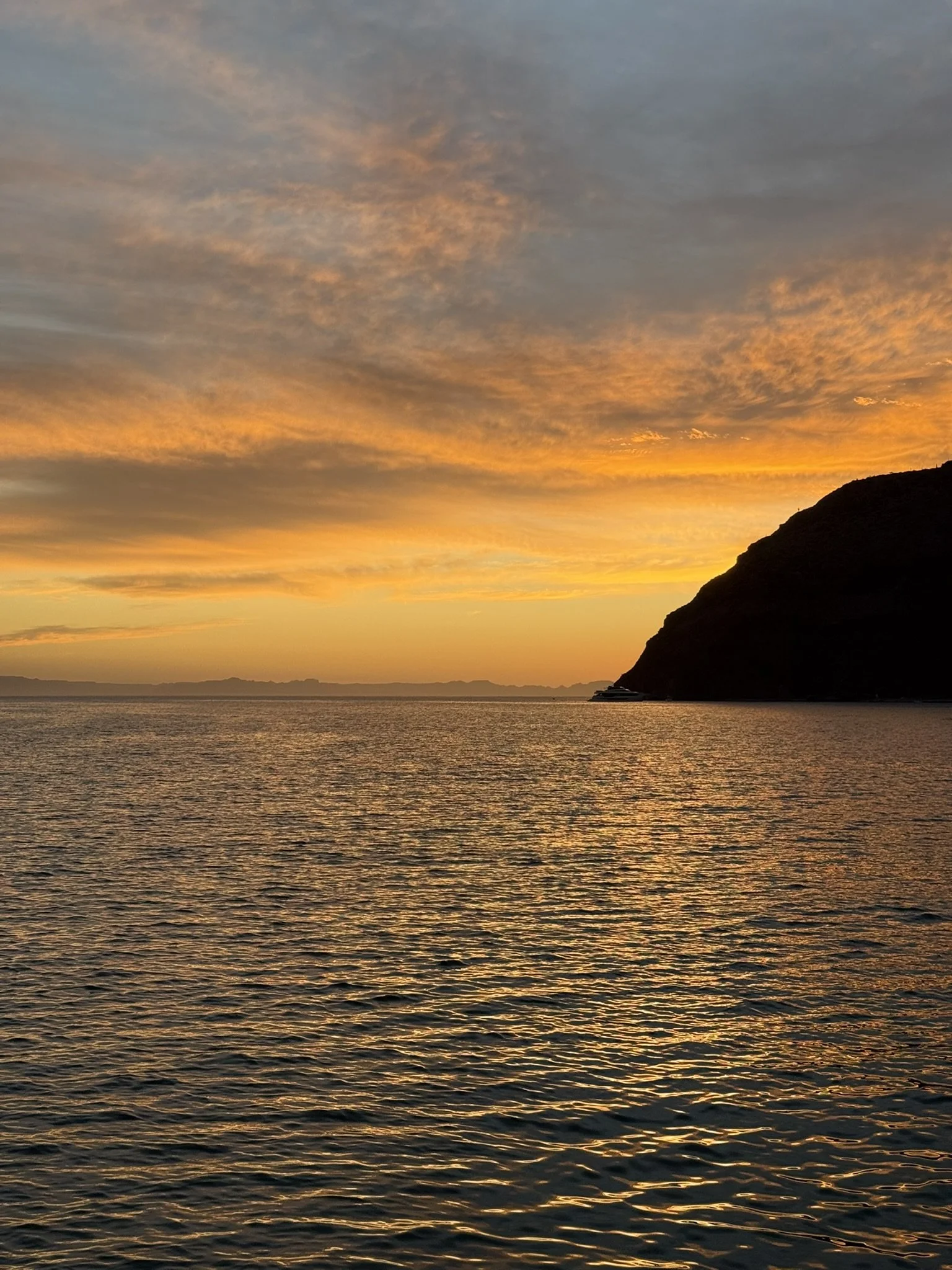 A sunset over the ocean with orange, yellow, and gray clouds in the sky and a dark silhouette of a hill or mountain on the right side.