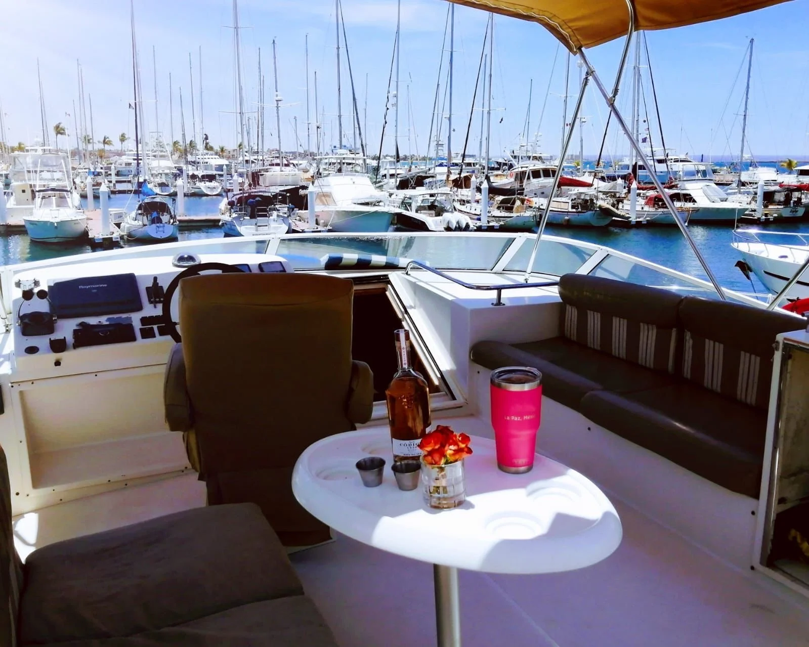 View from inside a boat showing a marina with numerous docked yachts and sailboats, and a table with drinks and decorations in the foreground.