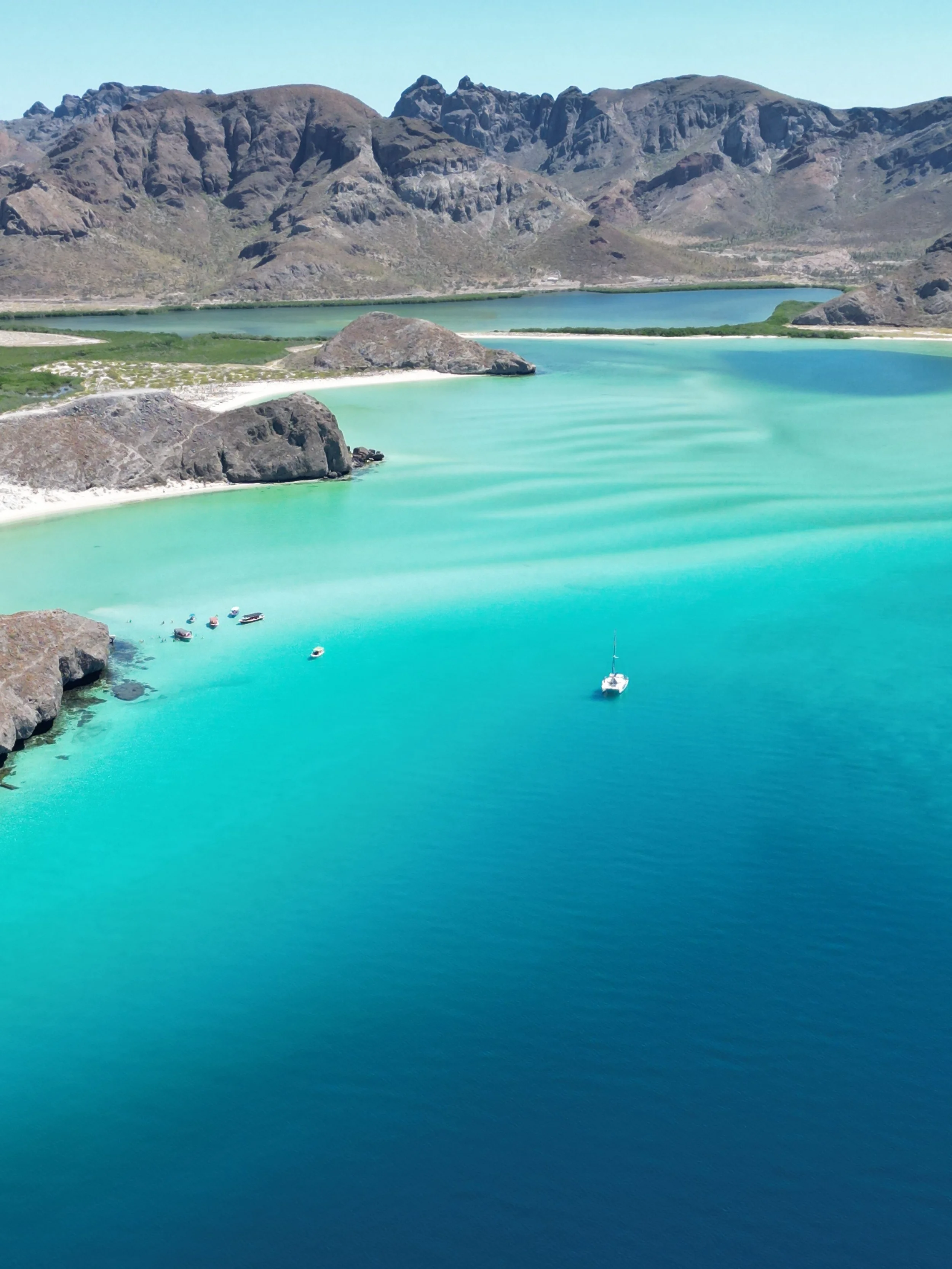 Aerial view of a turquoise bay with a sailboat and several smaller boats, surrounded by rocky islands and mountainous terrain under a clear blue sky.