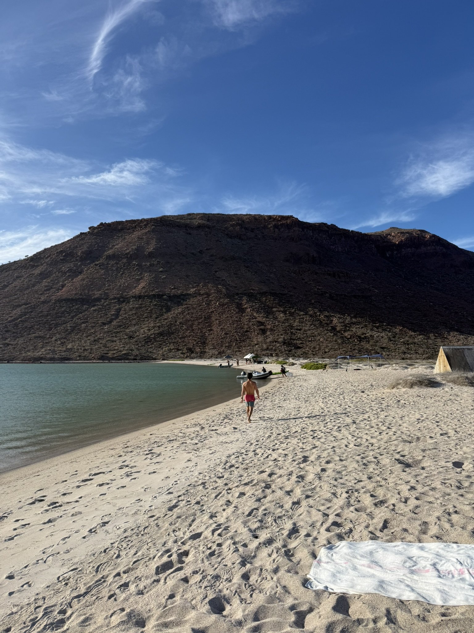 Person walking on sandy beach near water with a mountain in the background under a partly cloudy sky.