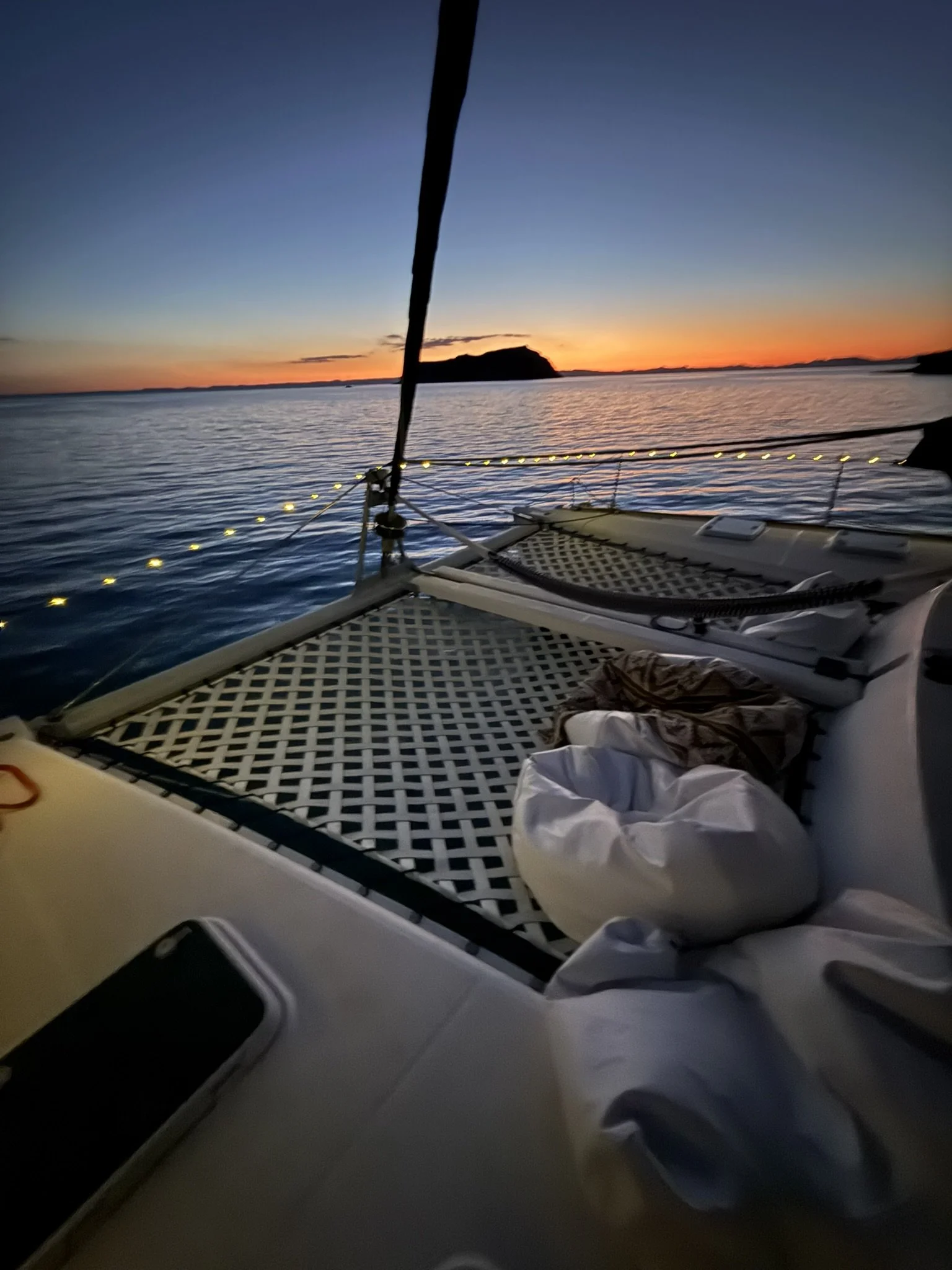 View from a boat at sunset showing a body of water, distant landmass, and a colorful sky with shades of blue, orange, and yellow. Part of the boat's deck and some items are visible.