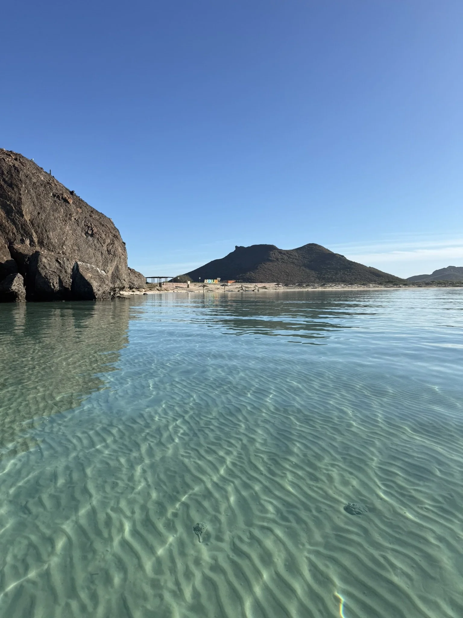 Clear water with ripples, large rocky formations on the left and a mountain in the background under a blue sky.