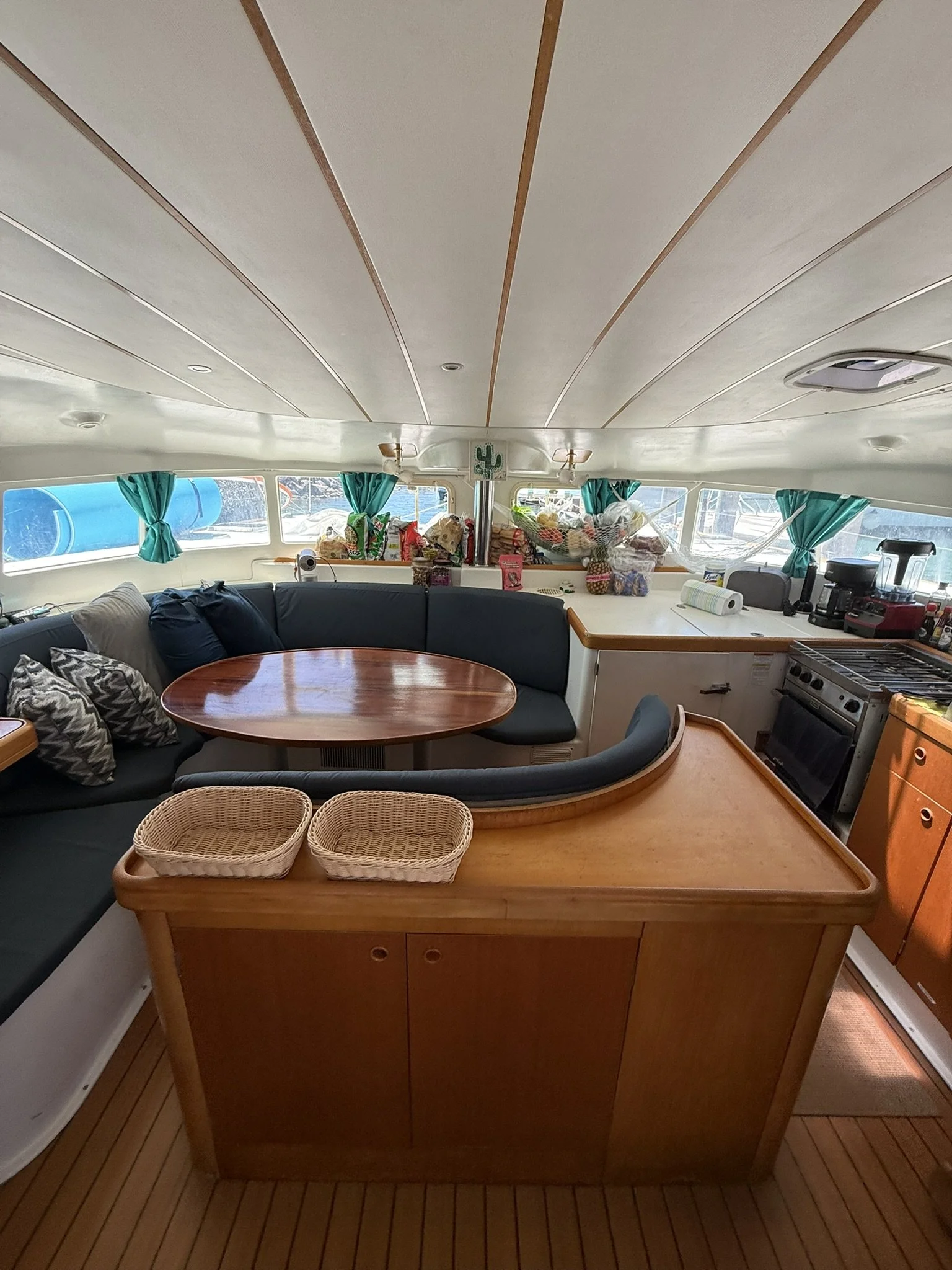 Interior of a boat or yacht with a curved seating area, wooden table, kitchen appliances, and multiple small baskets on a wooden countertop. Green curtains are covering the windows with a view of the water and boats outside.