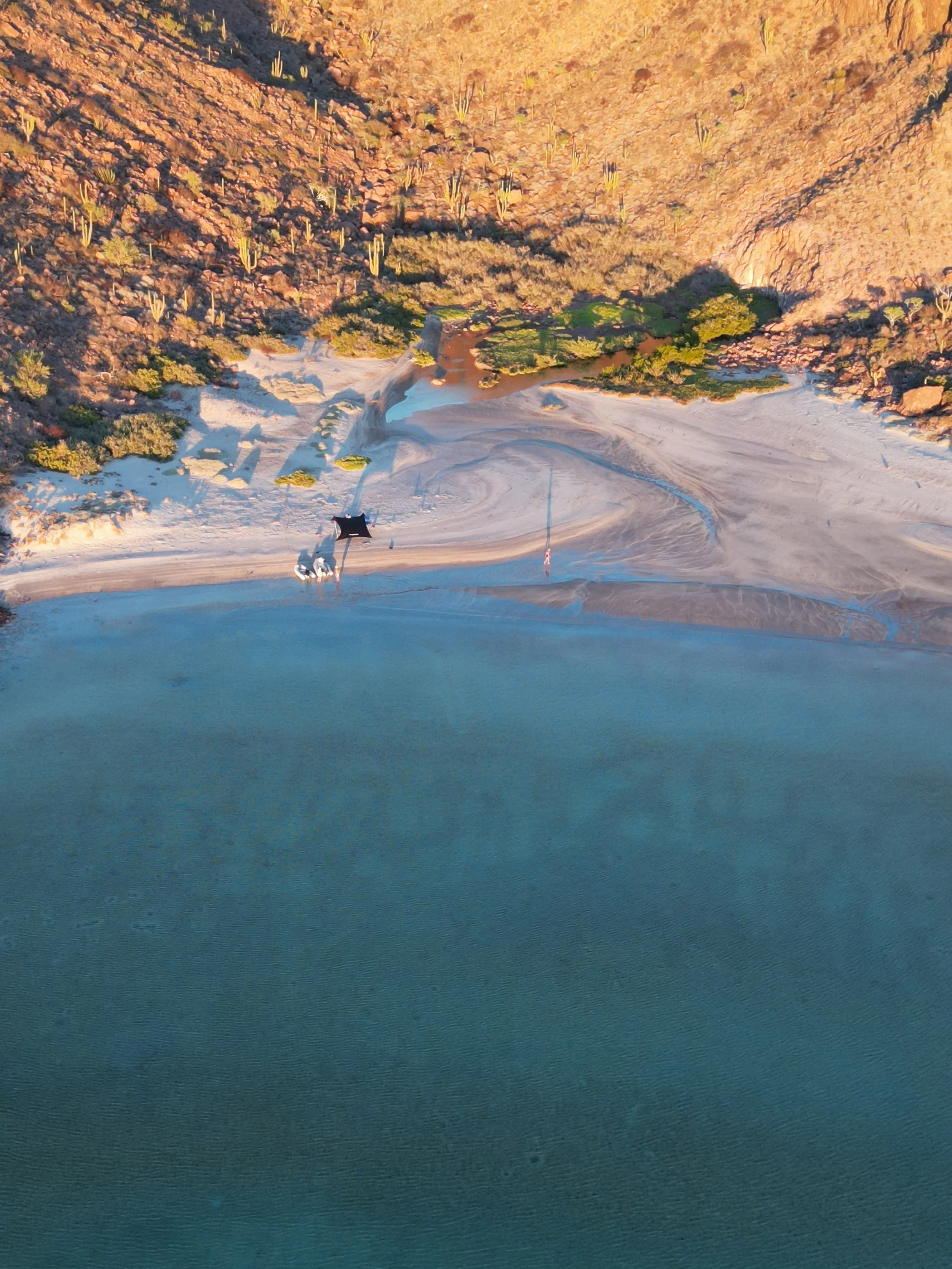 An aerial view of a lake with a sandy beach, surrounded by a rocky hillside with desert vegetation, seen in warm lighting.