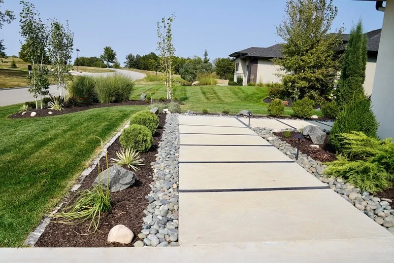 Concrete driveway with black lines, surrounded by landscaped yard with bushes, rocks, grass, and trees in front of a house.