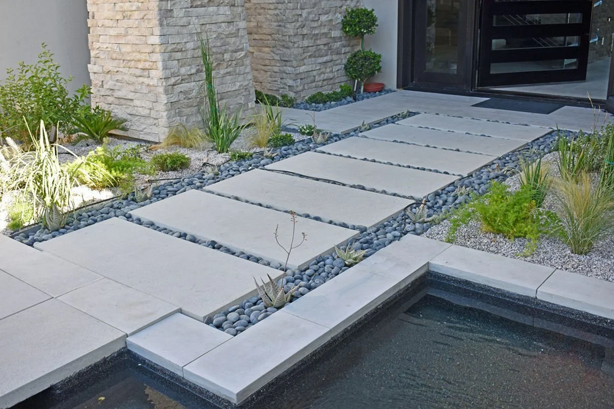 Modern pathway with large concrete slabs surrounded by small black pebbles, leading to a house entrance with stone walls and landscaped garden with various plants.