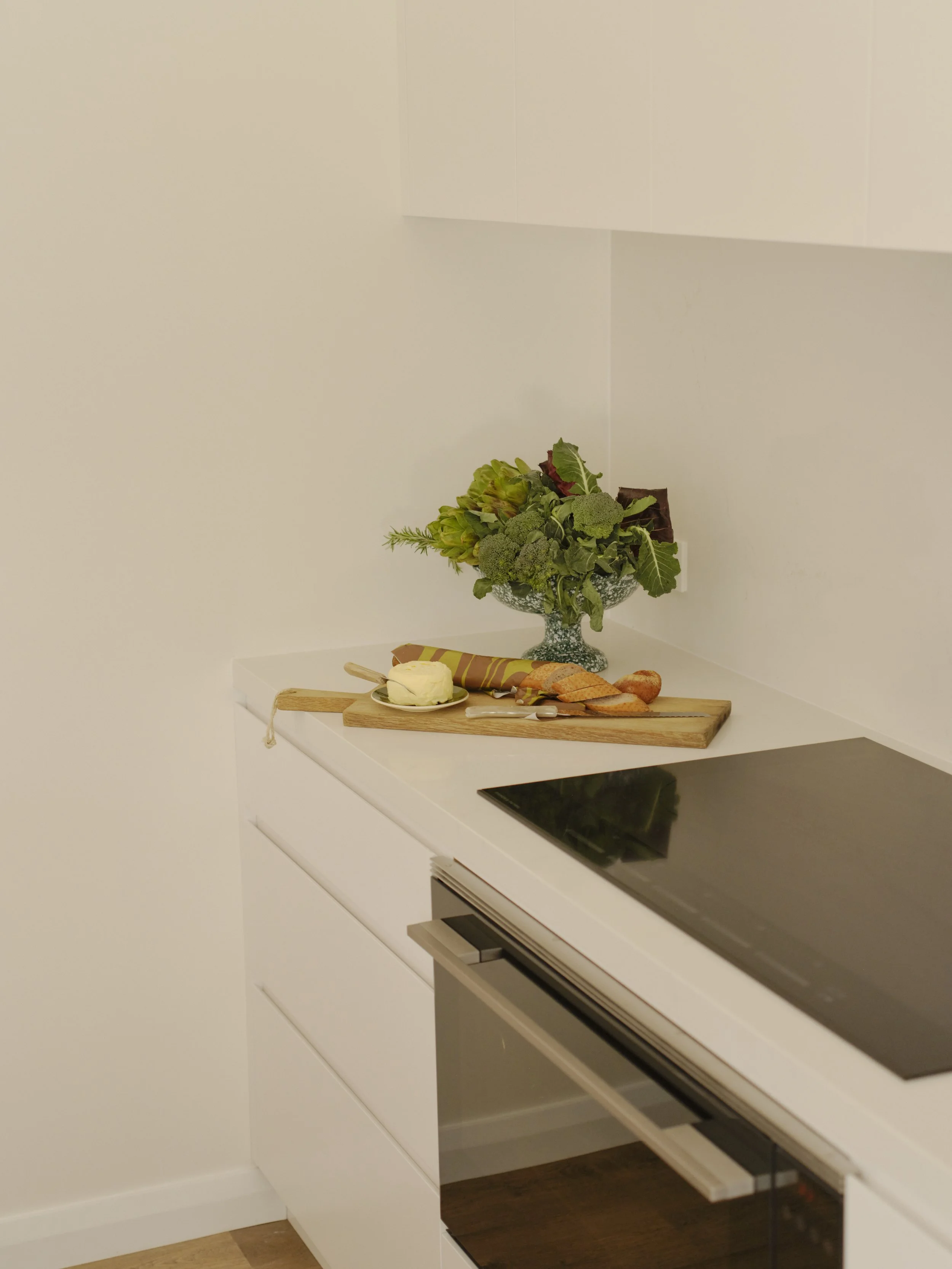 Kitchen countertop with a wooden cutting board, slices of bread, a knife, a small round cheese, and a decorative green leafy vegetable arrangement in a glass vase.