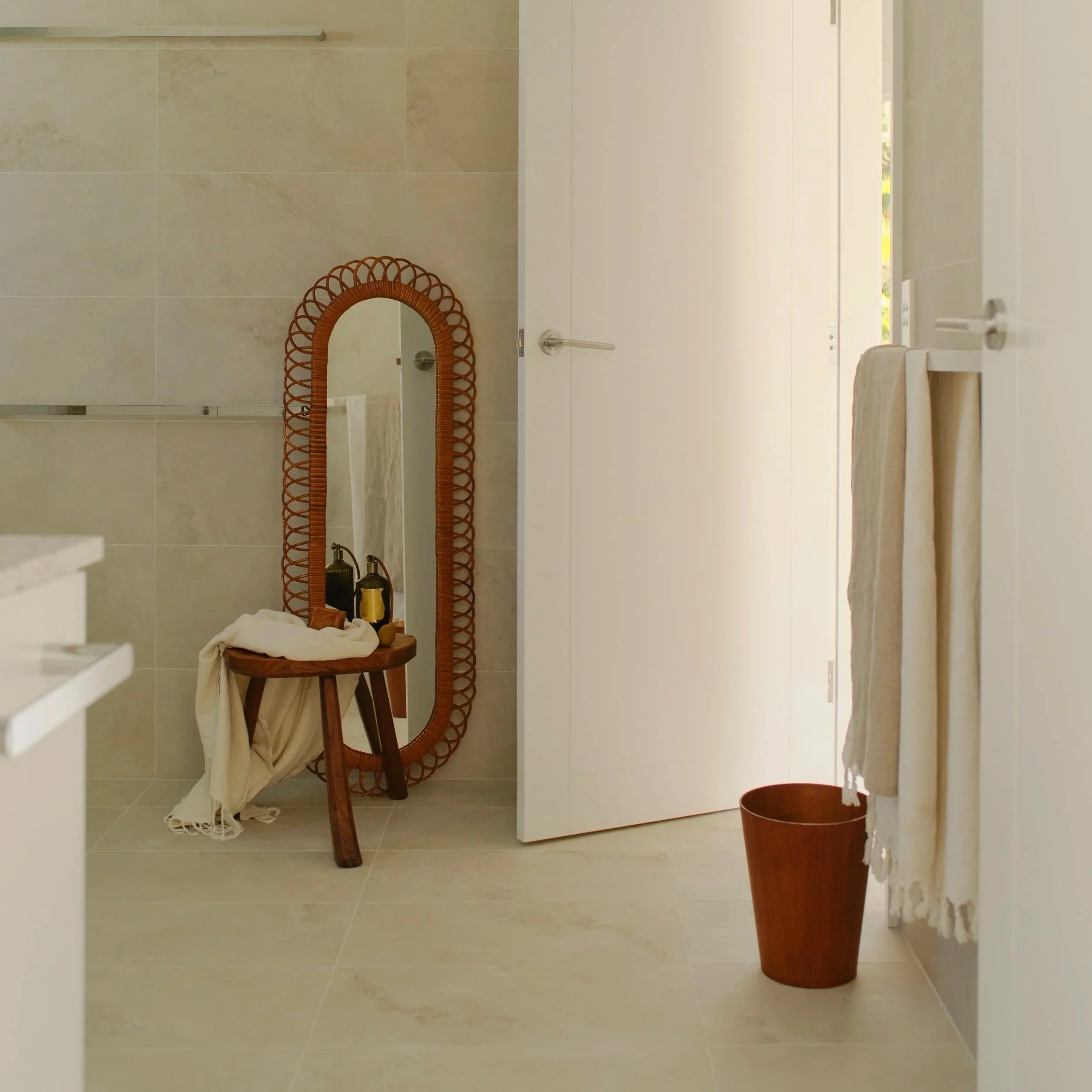 A bathroom corner with a tall, oval mirror in a brown wicker frame, a small wooden stool holding toiletries and a towel, a white door partially open, a beige towel hanging on a wall hook, and a brown trash can on the floor.