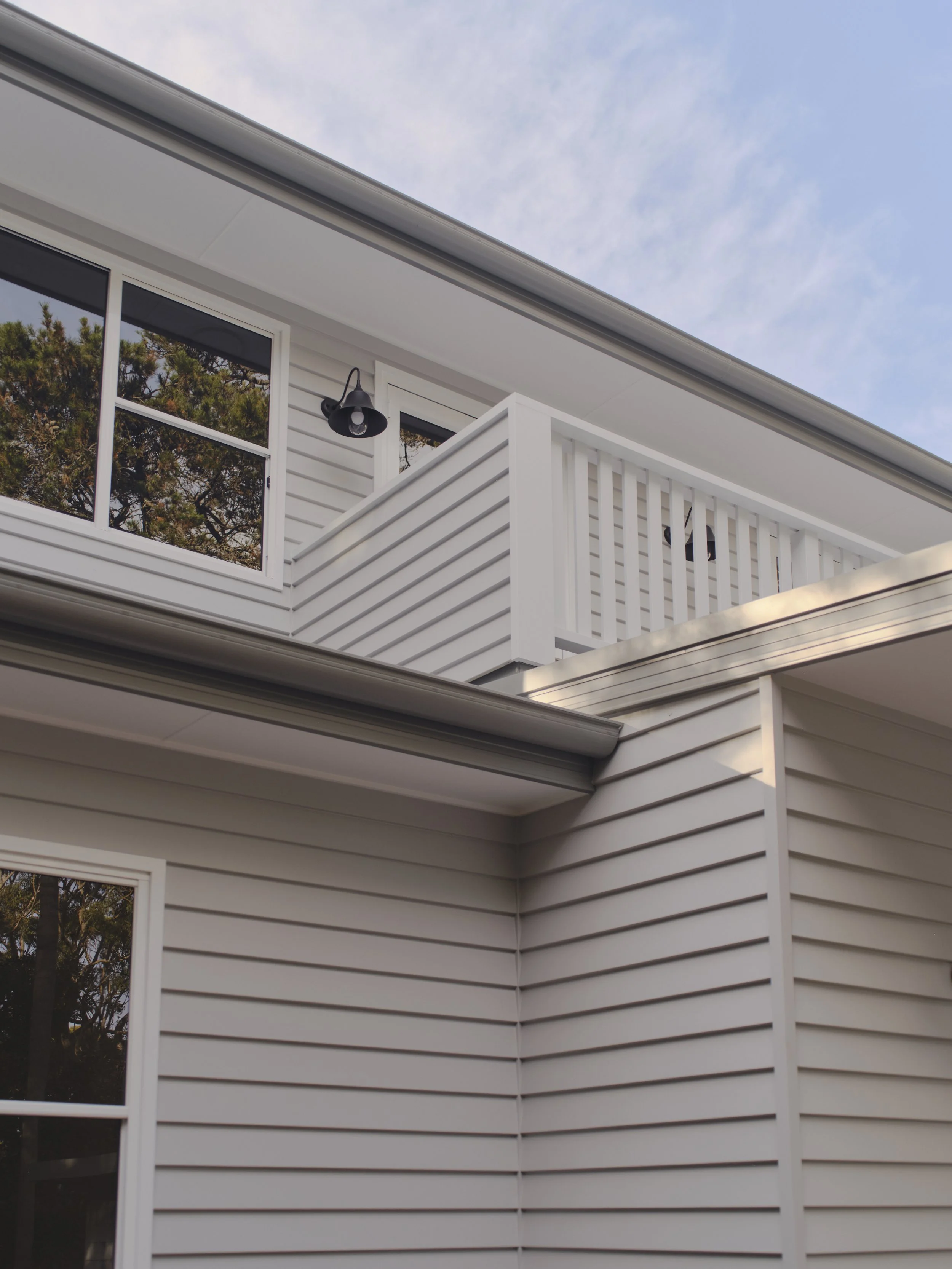 View of the exterior corner of a modern house with white siding, a balcony with white railing, a black outdoor wall light, and a window reflecting trees, under a partly cloudy sky.