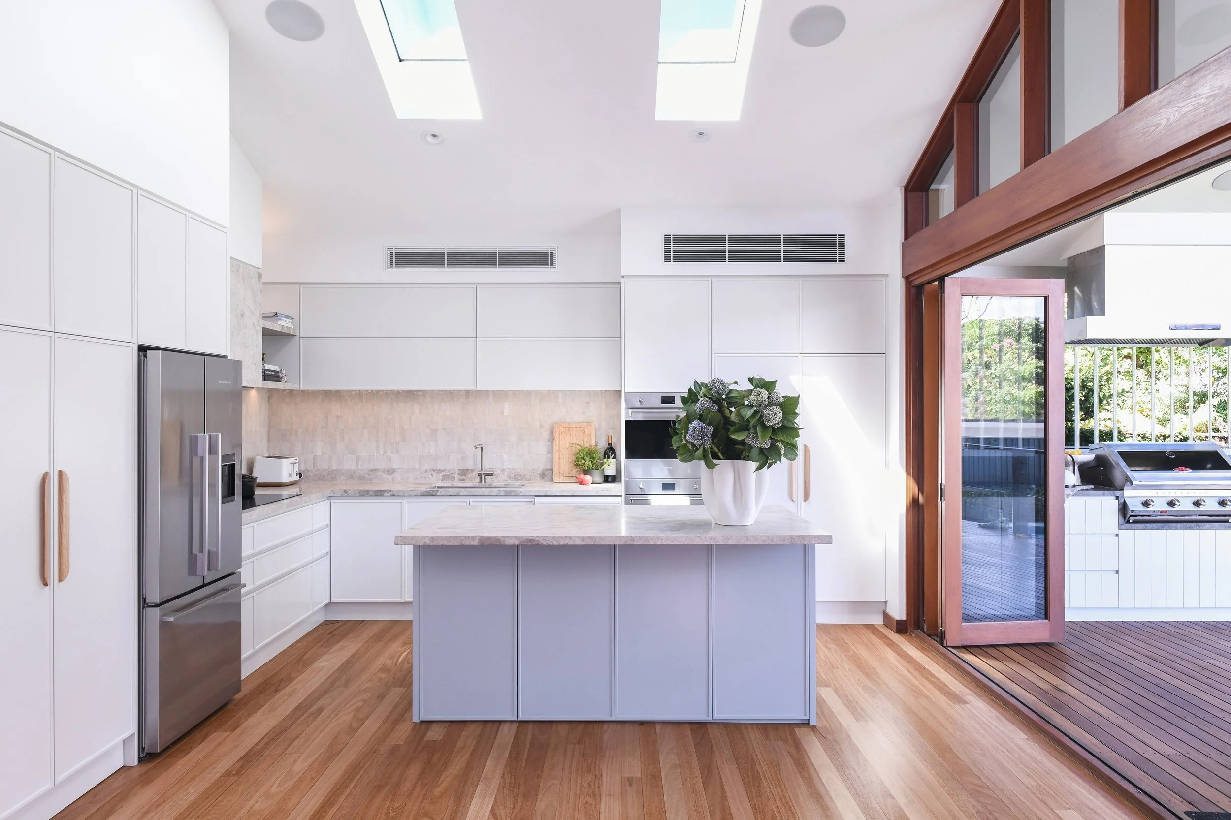 Modern kitchen with white cabinets, marble island, wooden flooring, skylights, potted plant on island, and sliding glass door leading to outdoor deck with grill.