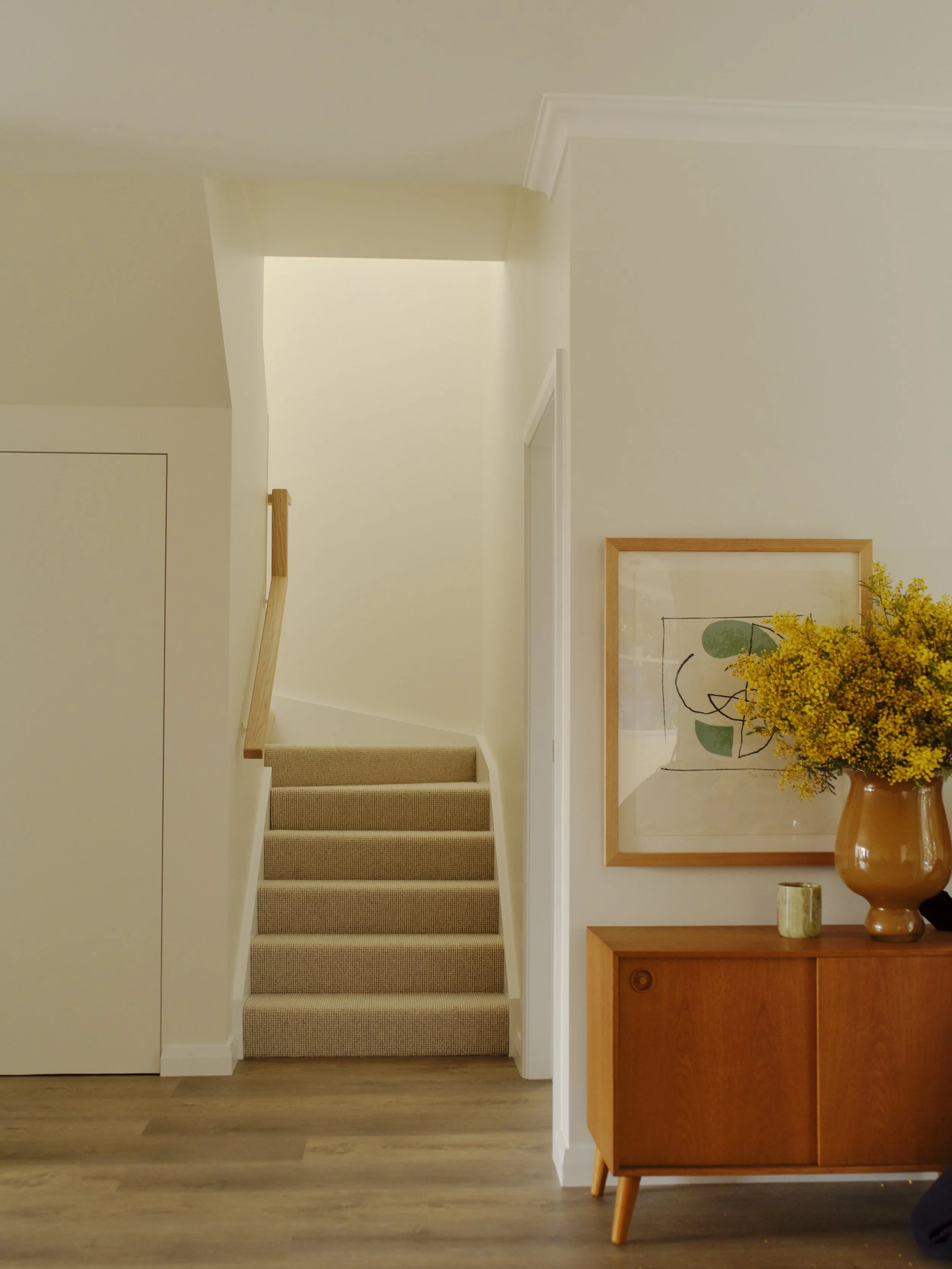 Interior view of a hallway with a staircase leading upward, a framed abstract art piece, a vase with yellow flowers, and a small decorative item on a wooden sideboard.