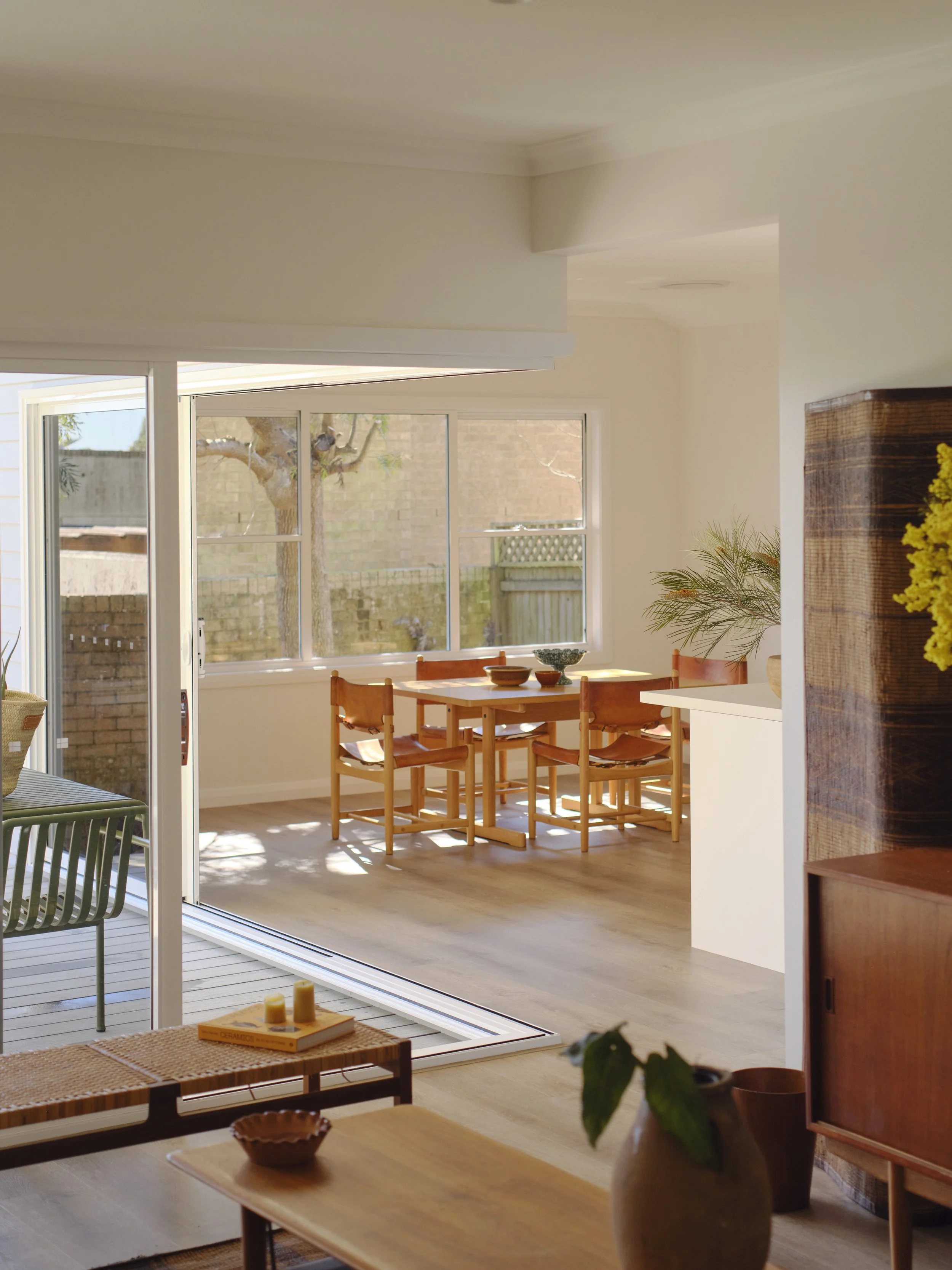 Bright dining room with wooden table and chairs, large window, sliding glass door leading to outdoor patio, and decorative plants.