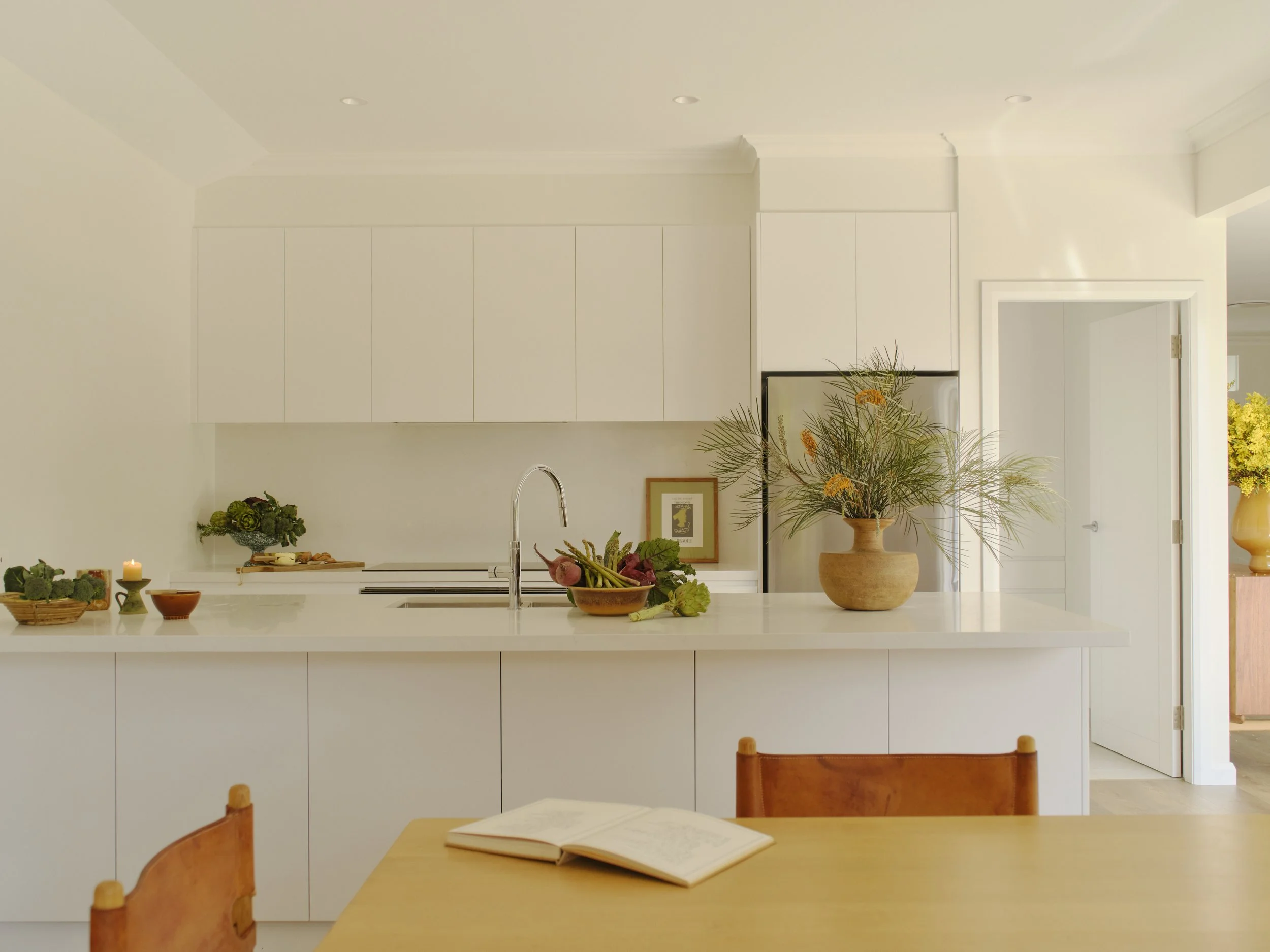Bright, modern white kitchen with minimalist design, decorated with potted plants, an open book on a wooden table, and a vase with greenery on the counter.