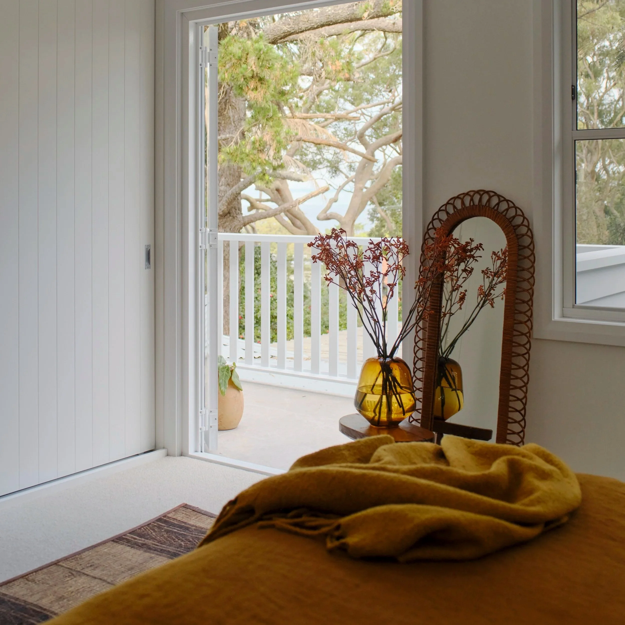 Interior of a bedroom with a view of the balcony outside, showing a tree with twisted branches. The room has a bed with a yellow blanket, a mirror, and two yellow vases with dried branches.