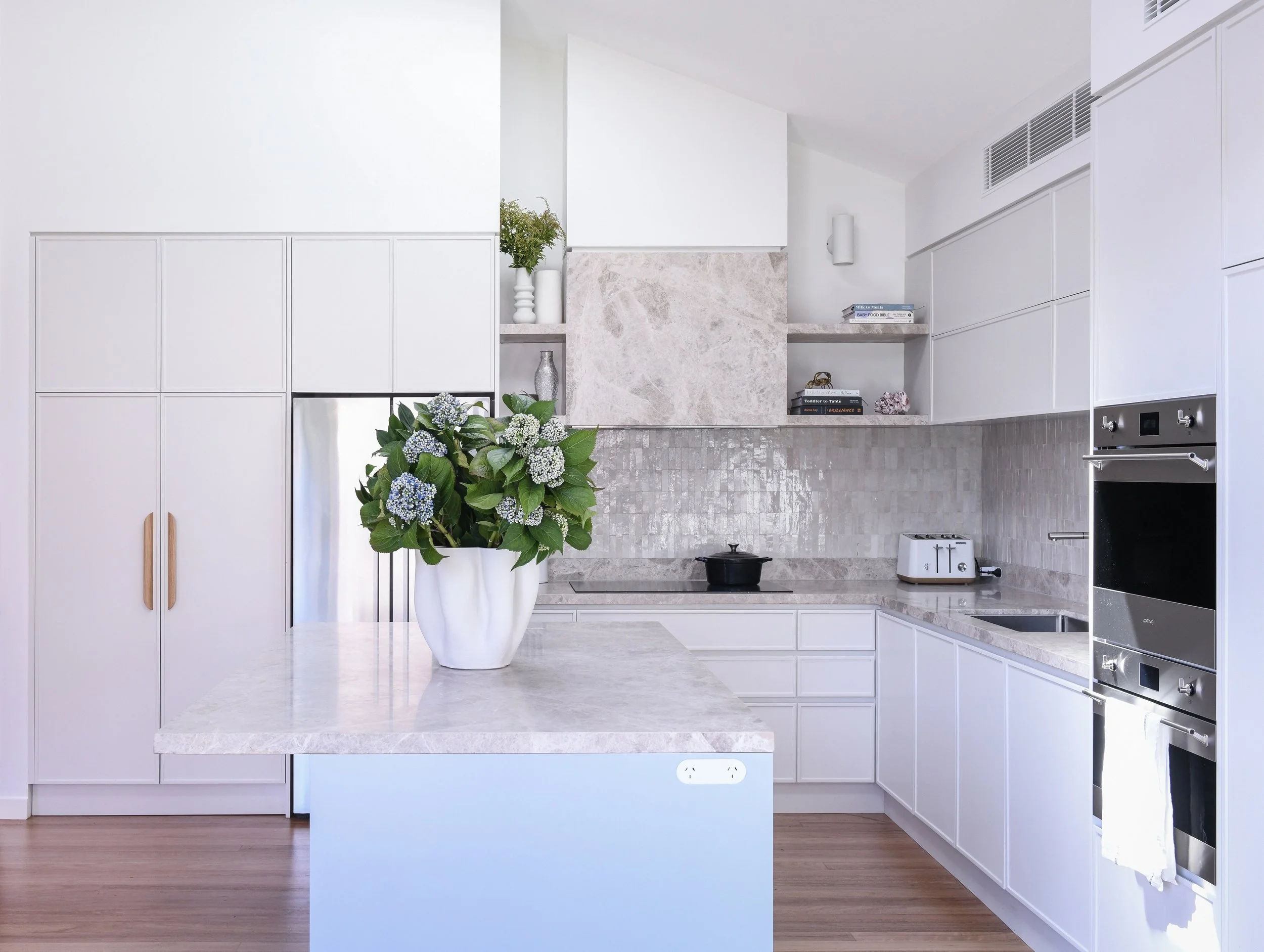 Minimalist white kitchen with marble countertops, backsplash, and built-in appliances, decorated with a large potted flower on the island counter.