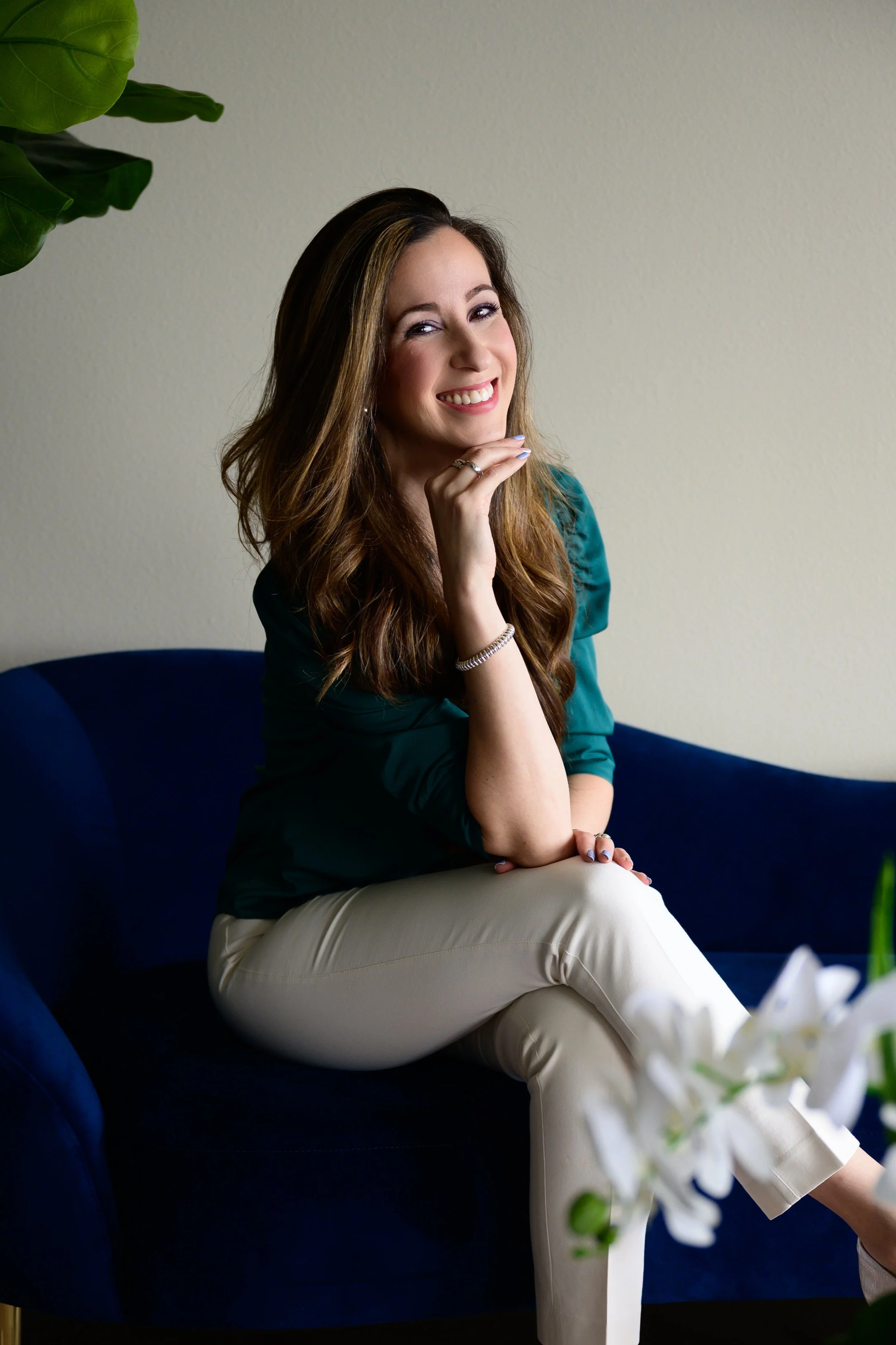 A smiling woman with long brown hair, wearing a teal blouse and beige pants, sitting on a dark blue sofa, touching her chin with her right hand, and looking at the camera.