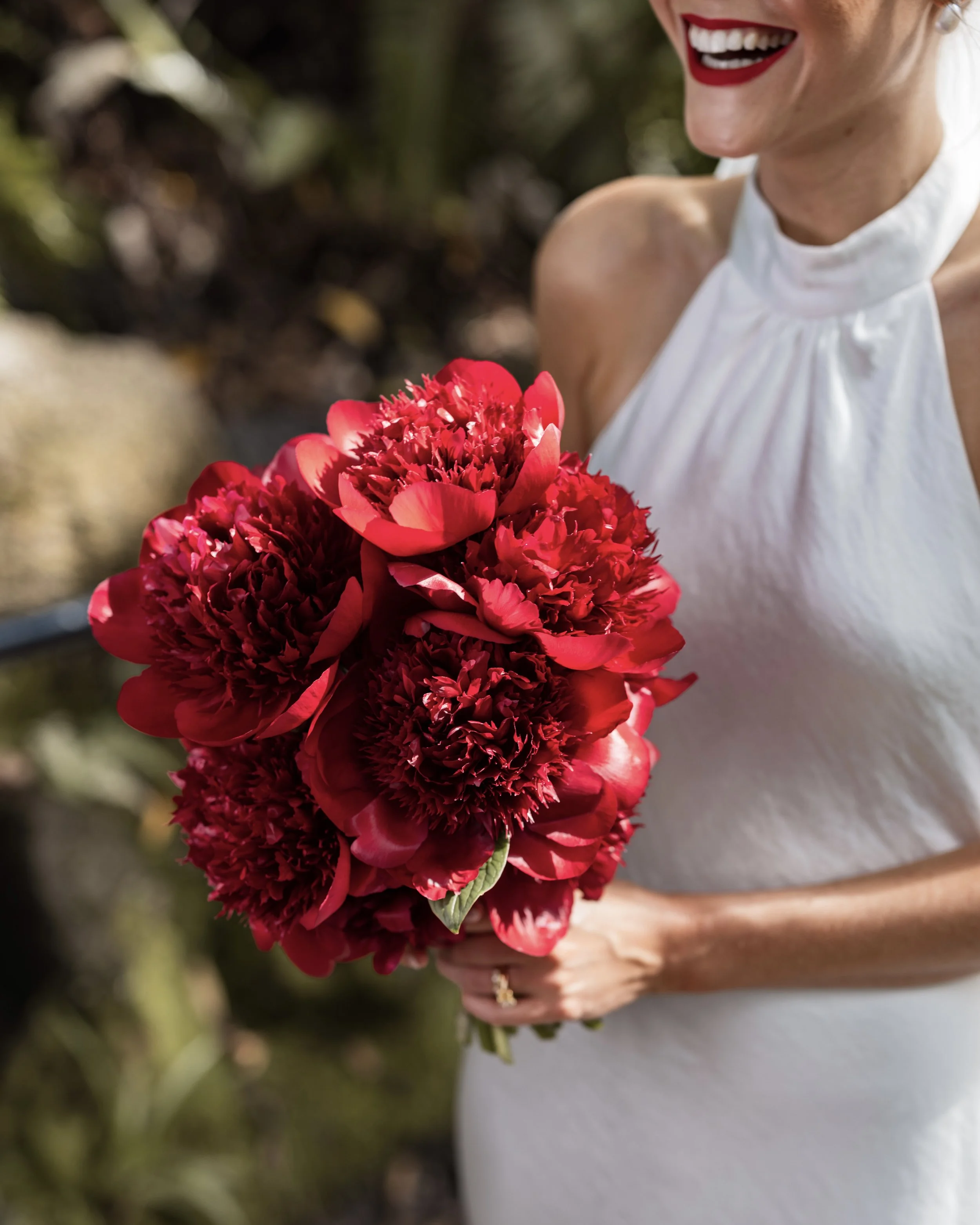 A woman wearing red lipstick in a wedding dress holding a bridal bouquet of large red peony flowers.