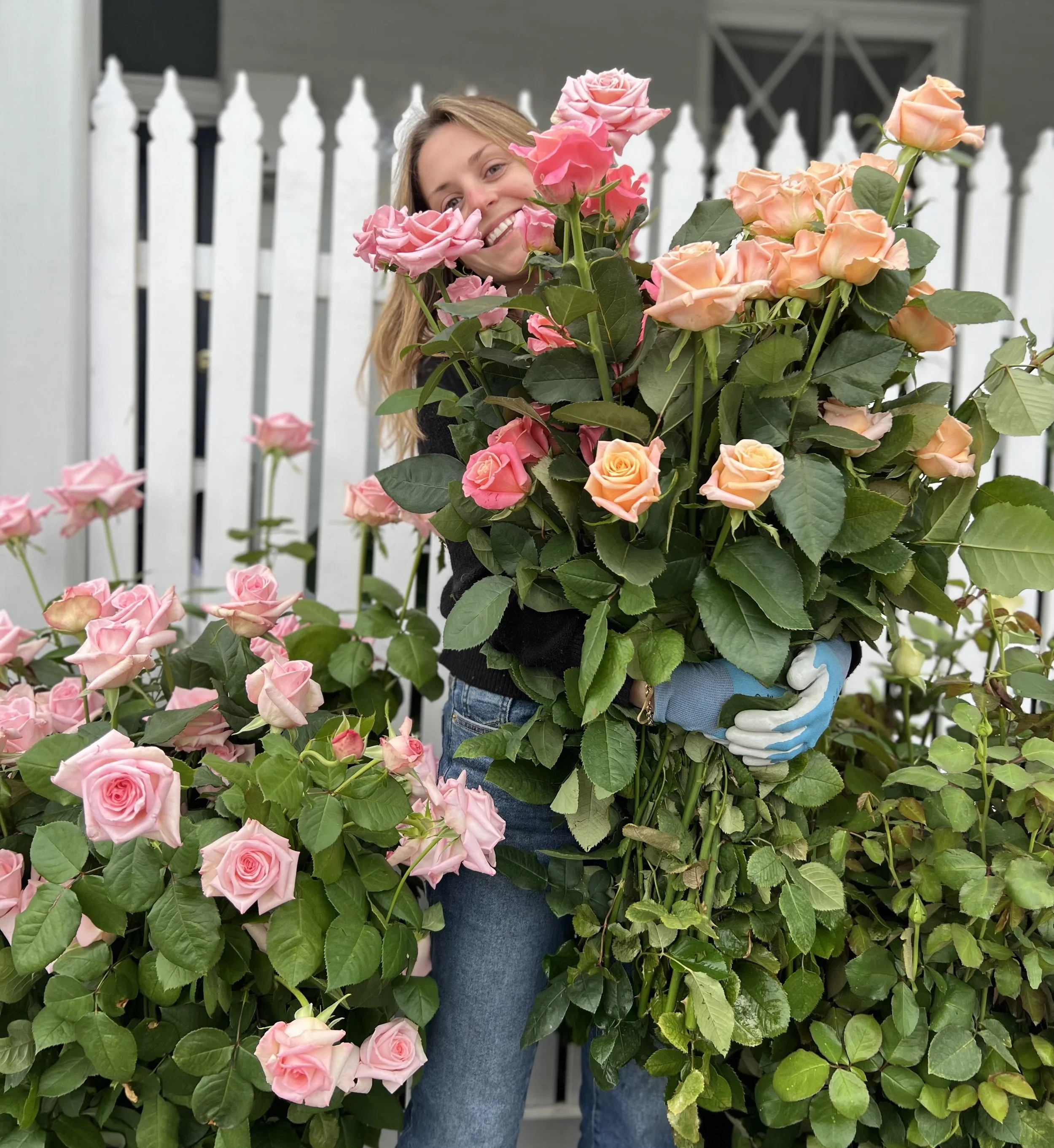 A woman smiling while holding tall, wild stems of pink and orange roses with a white picket fence in the background.