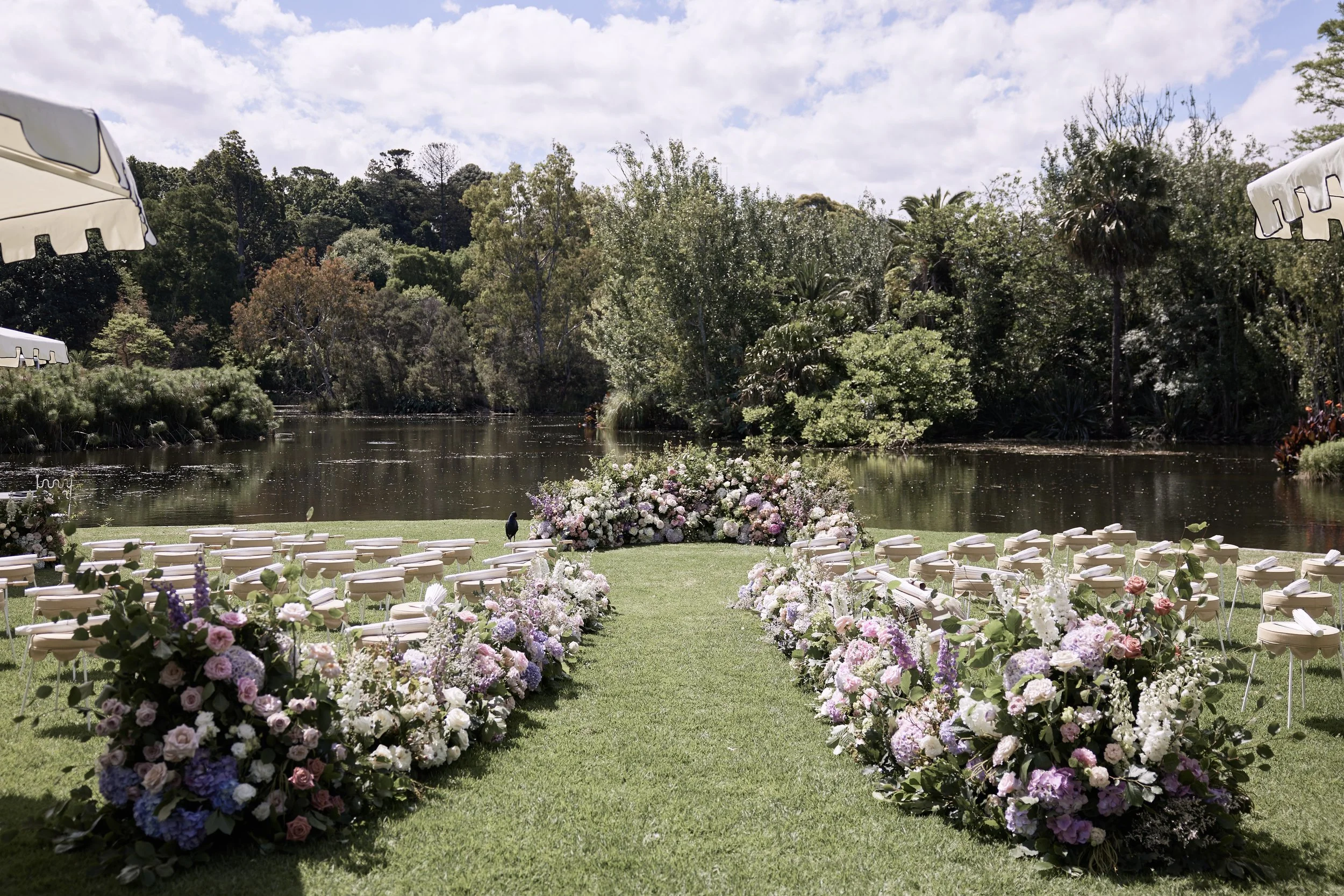 Outdoor wedding setup, featuring aisle adorned with voluminous floral arrangements featuring premium roses and hydrangeas. Accompanied by white chairs, a pond, and lush green trees under a partly cloudy sky.