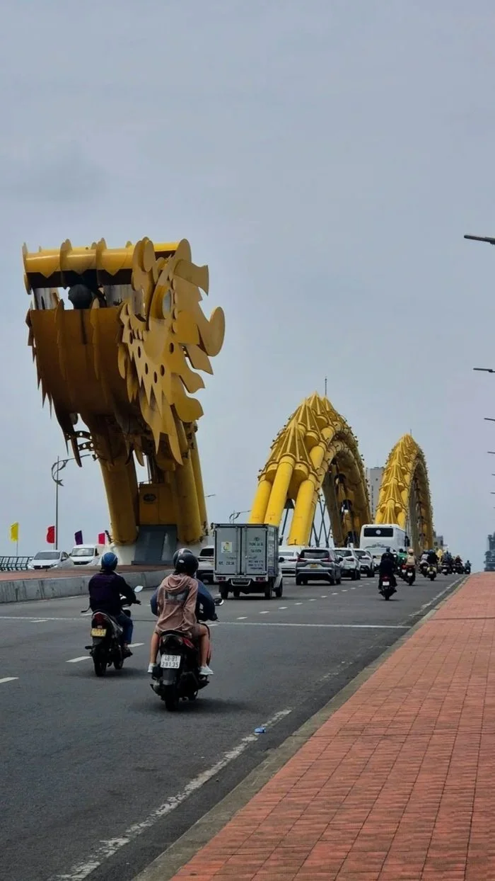 A bridge with large, yellow, dragon head sculptures as decorative arches on a busy road with motorcycles, cars, and a bus passing underneath.