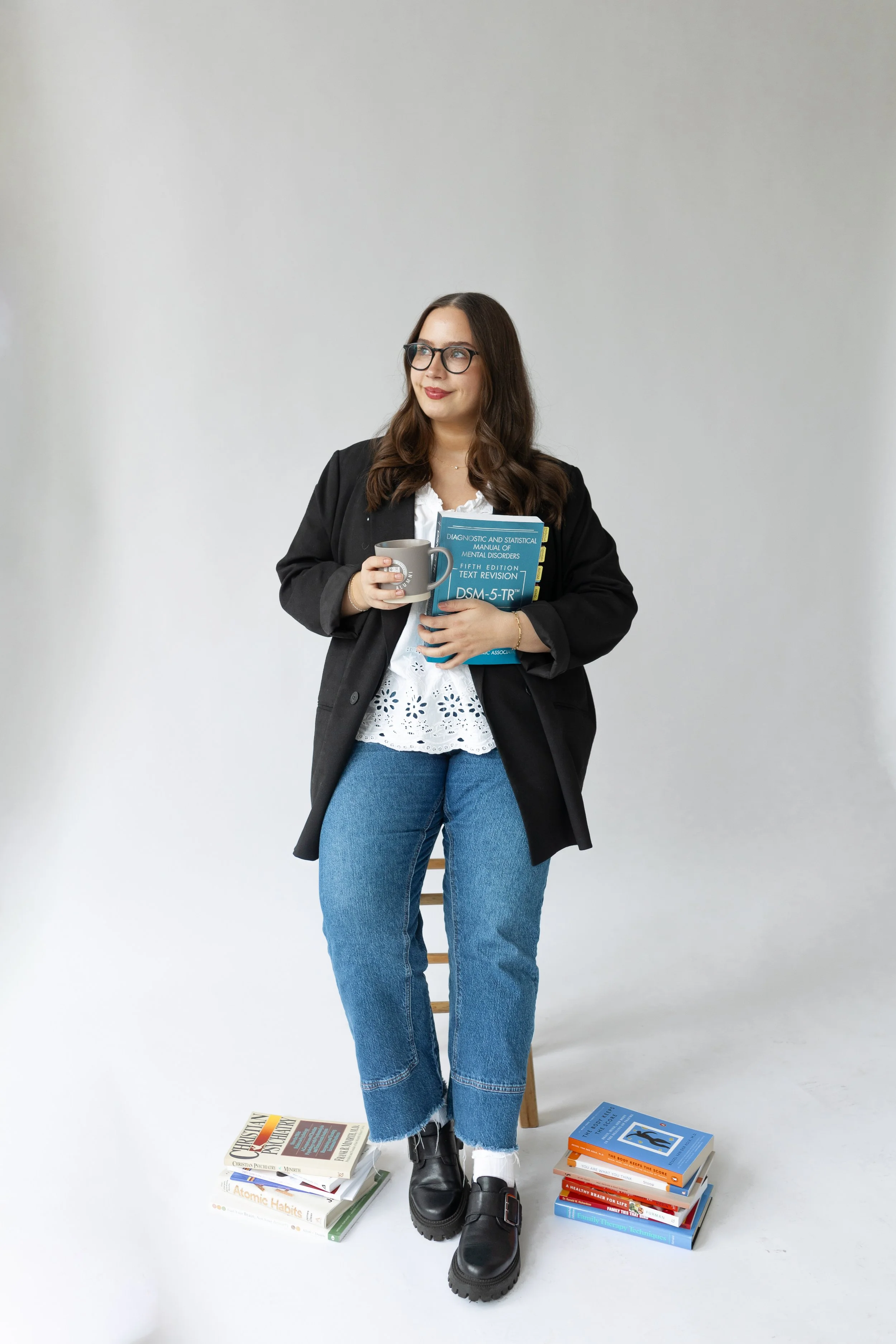 A woman with dark hair and glasses stands in front of a plain white wall, holding a mug and a book. She is dressed in a white lace top, jeans, black boots, and a black blazer. Books are stacked on the floor and a chair, some on the floor and some on the chair.