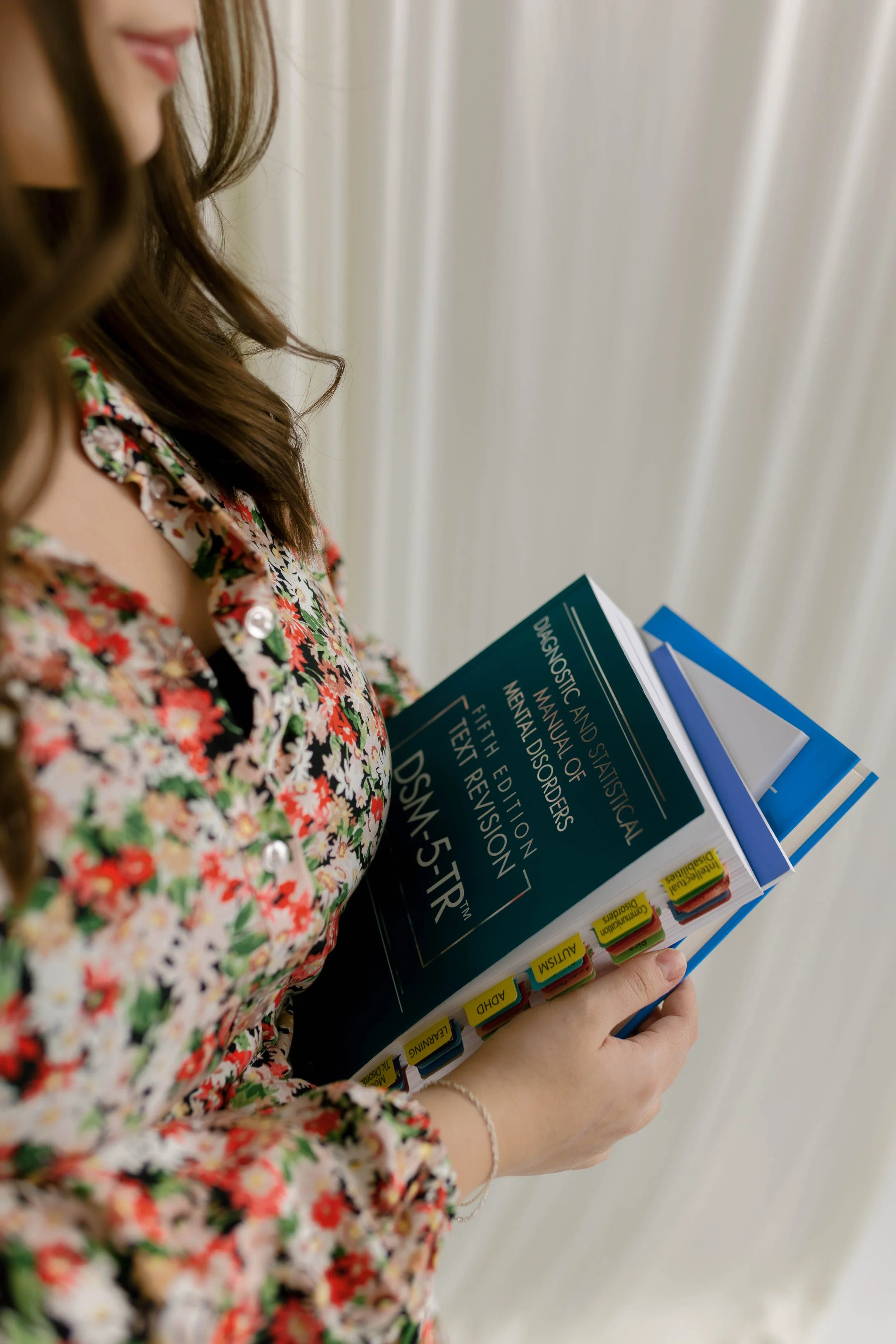 A person with long brown hair, wearing a floral shirt, holding a stack of medical textbooks and binders, with visible tabs labeled 'AUSTISM,' 'LEARNING,' 'ADHD,' 'TEXT REVISION,' and 'MENTAL DISORDERS,' standing against a neutral background.