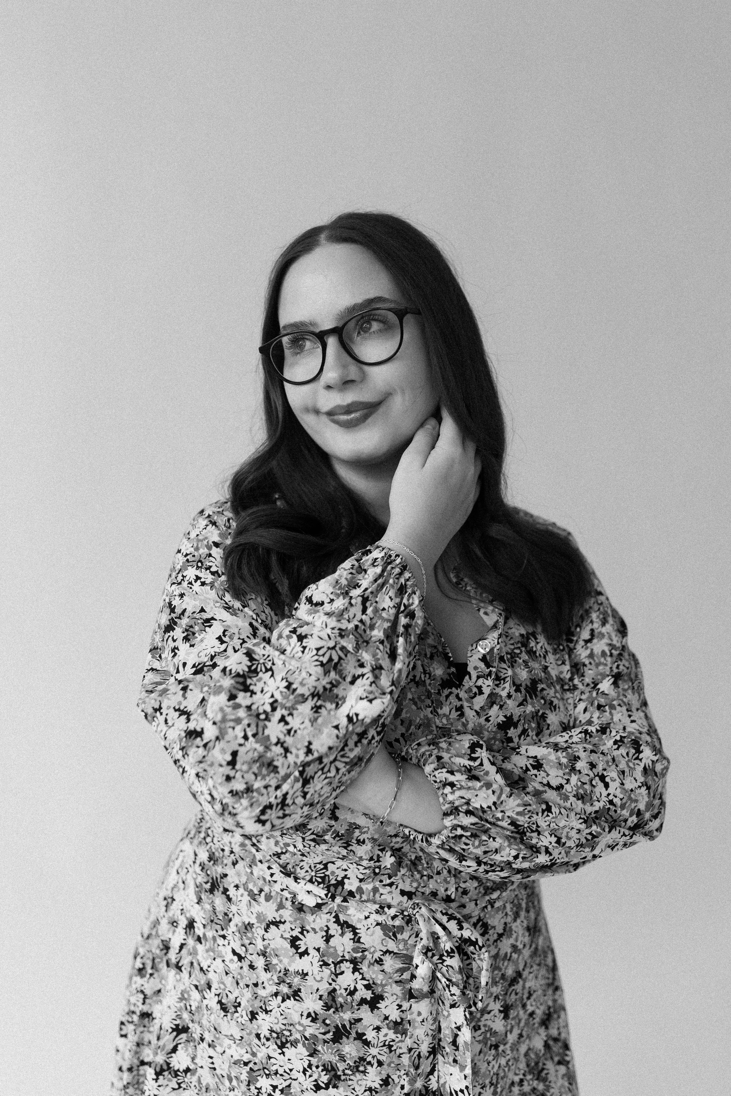 Black and white portrait of a woman with long dark hair, wearing glasses, a floral blouse, and a bracelet, posing with her hand near her neck and smiling.