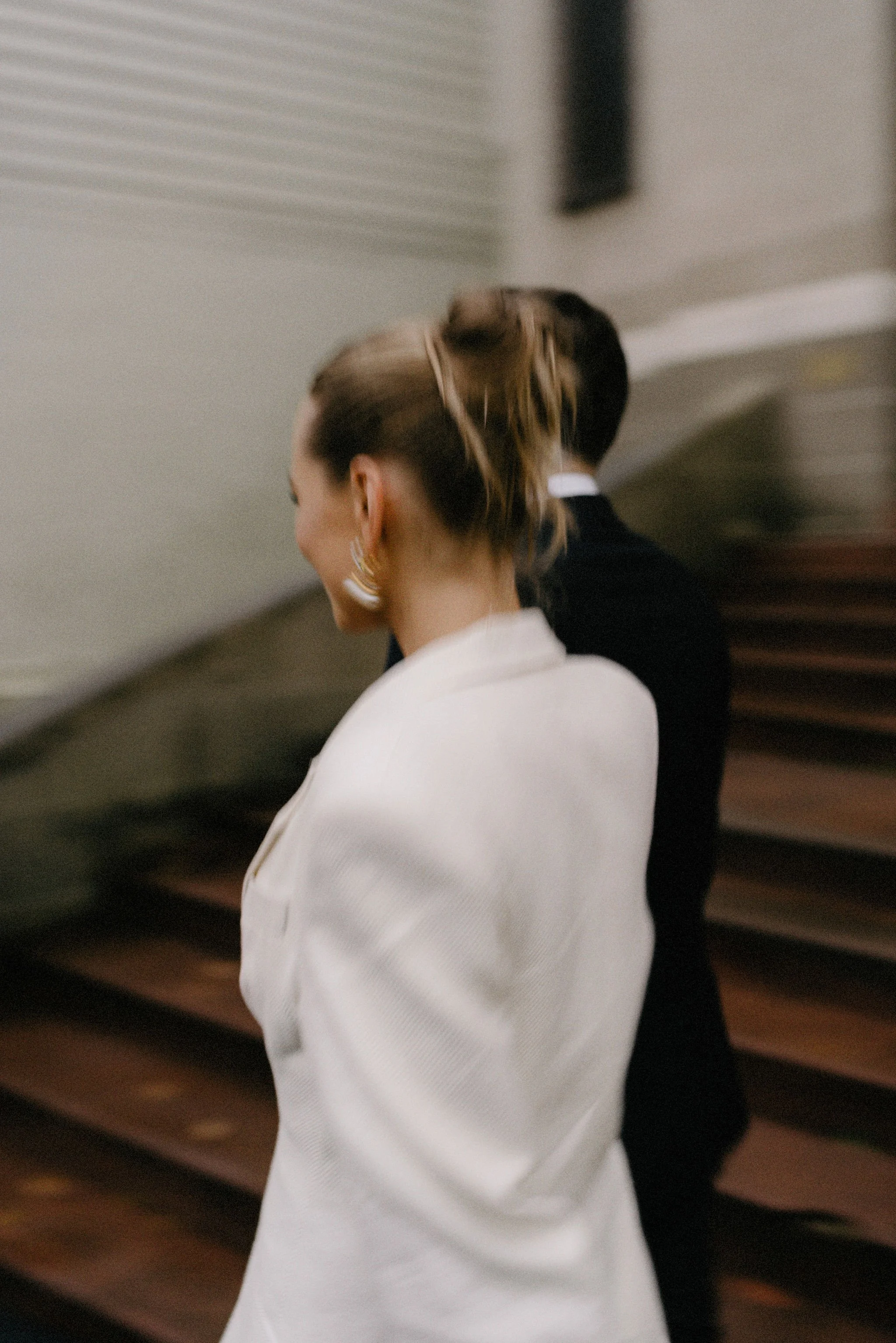 A woman with styled hair and large earrings, dressed in a white blazer, standing with a man in formal attire, blurry background with wooden stairs.