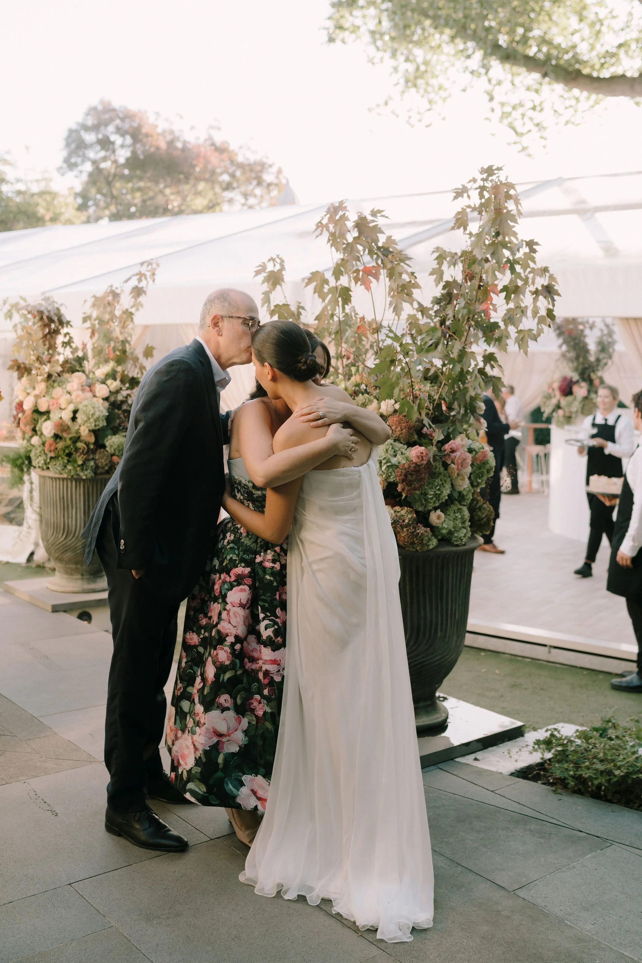 A embrace moment between a man, a woman in a black floral dress, and a woman in a white strapless wedding gown at an outdoor wedding reception with flowers.