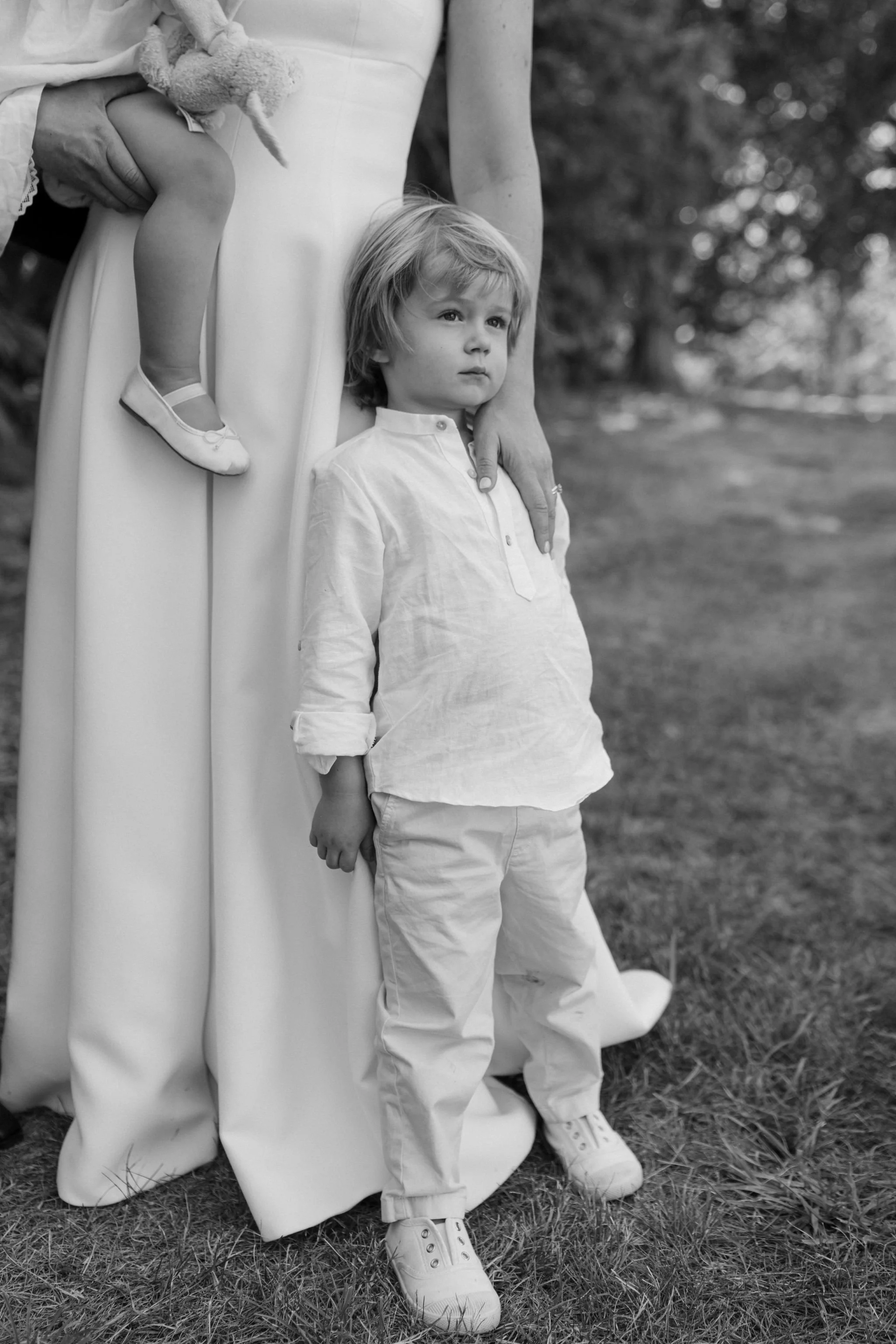 A woman standing outdoors next to a young boy, with a young girl sitting on her shoulder. The woman is wearing a long dress, and the boy has light-colored hair, a light shirt, and pants. The girl holds a stuffed bunny.