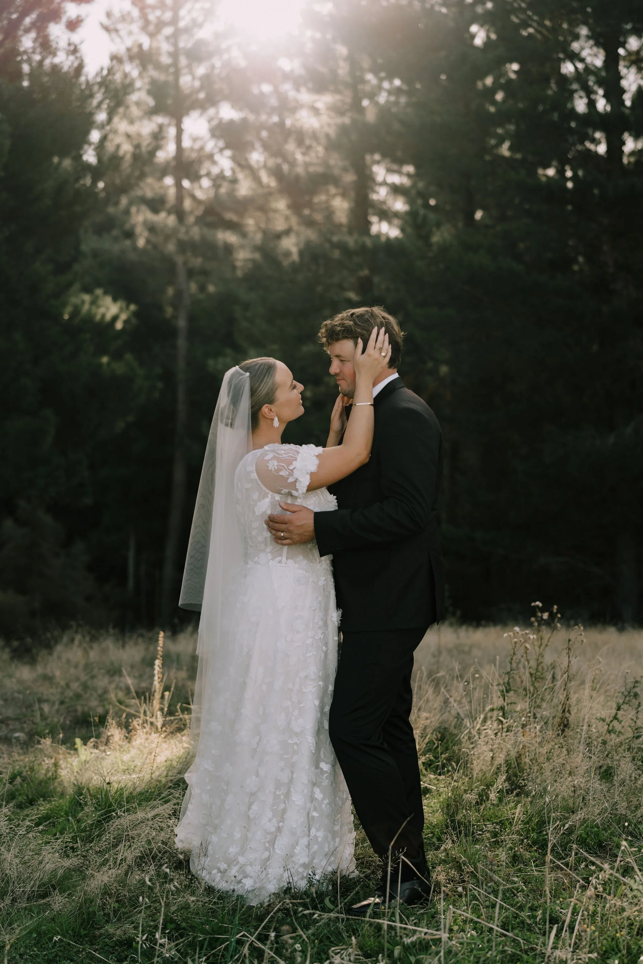 A bride and groom stand in an outdoor field, embracing and gazing into each other's eyes, with trees and sunlight in the background.
