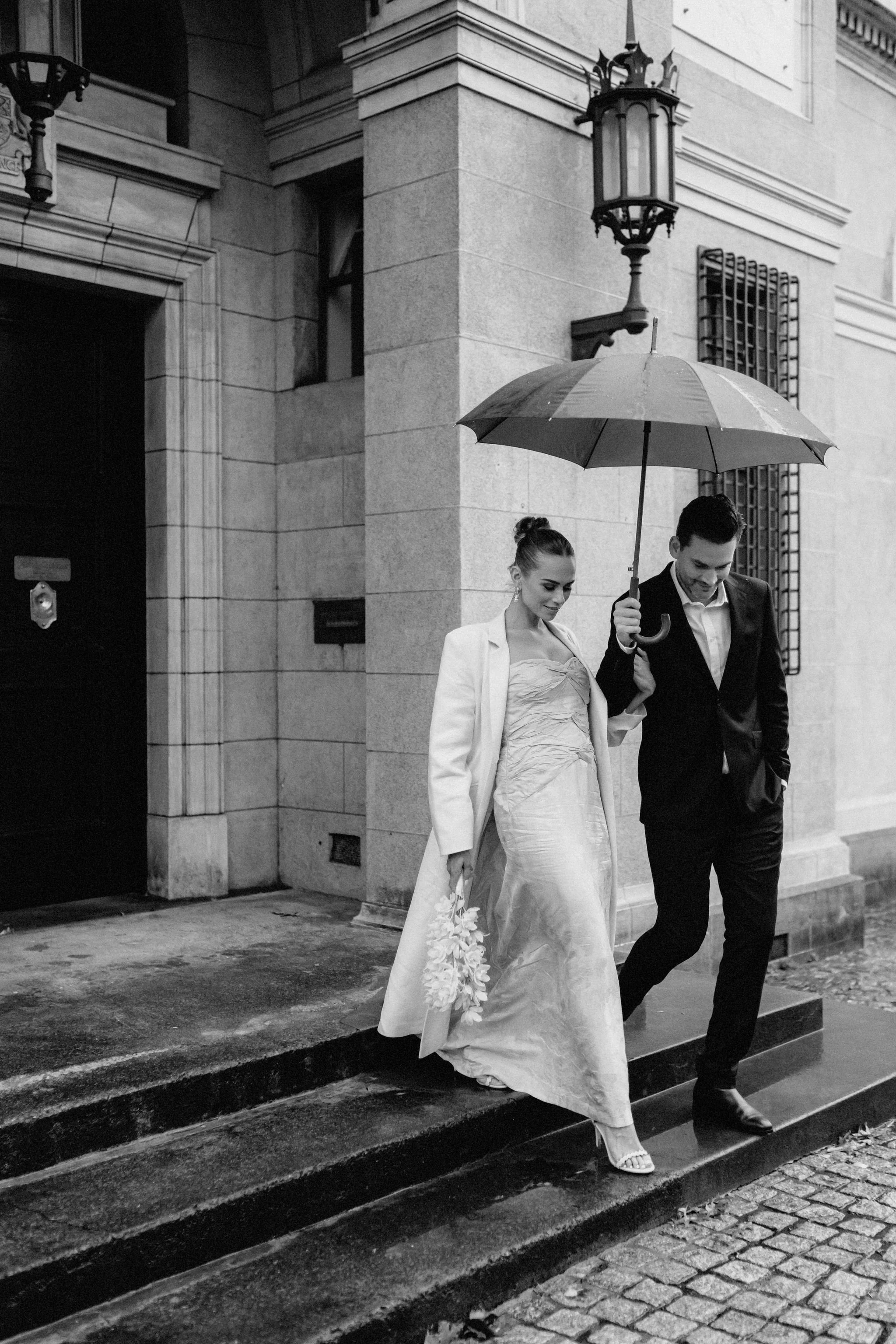 A black and white photo of a couple walking down the steps outside a building. The woman is wearing a long dress and holding a flower, while the man is dressed in a suit. They are sharing an umbrella on a rainy day.