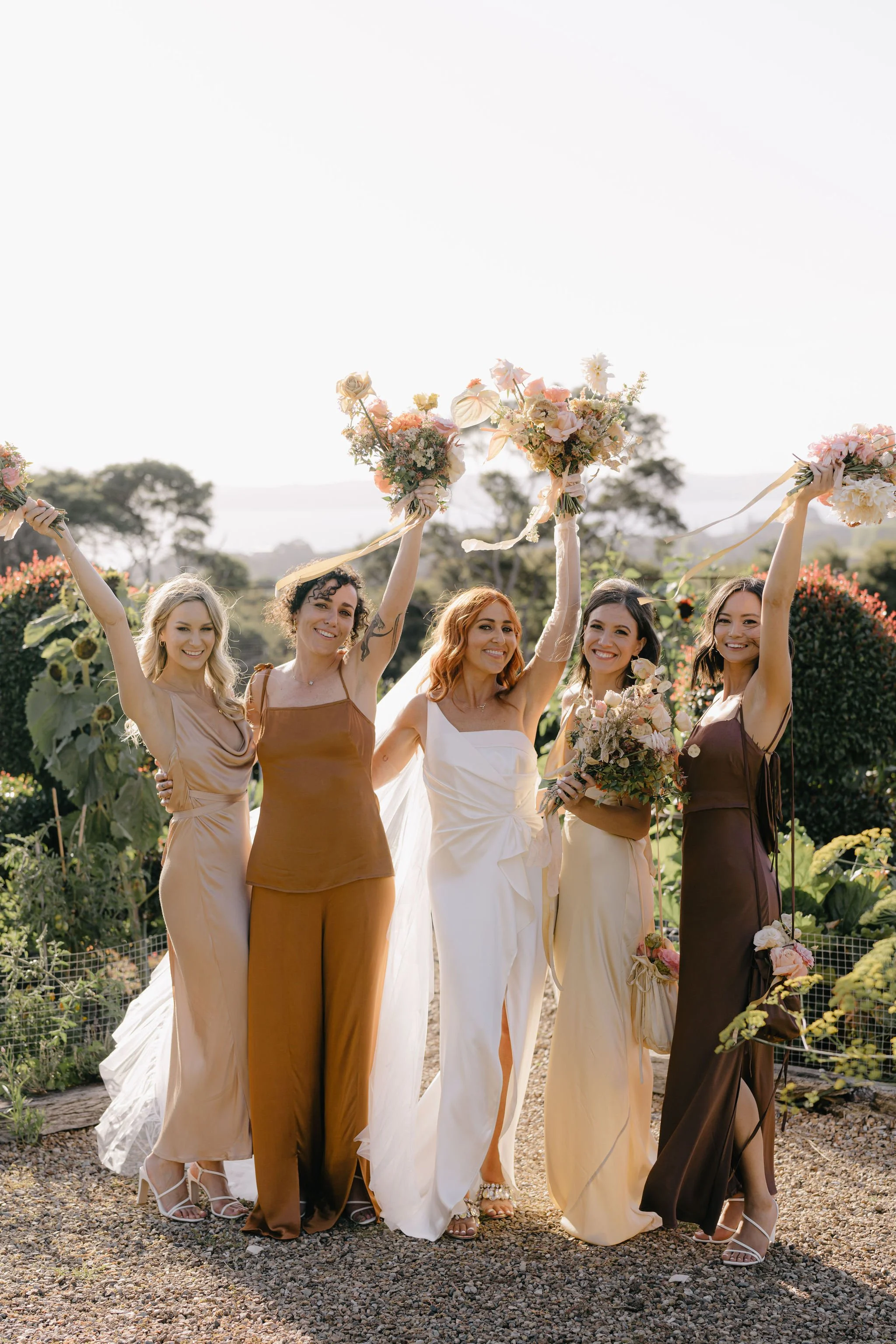 Group of five women celebrating outdoors, holding bouquets and raising their arms, dressed in colorful outfits.