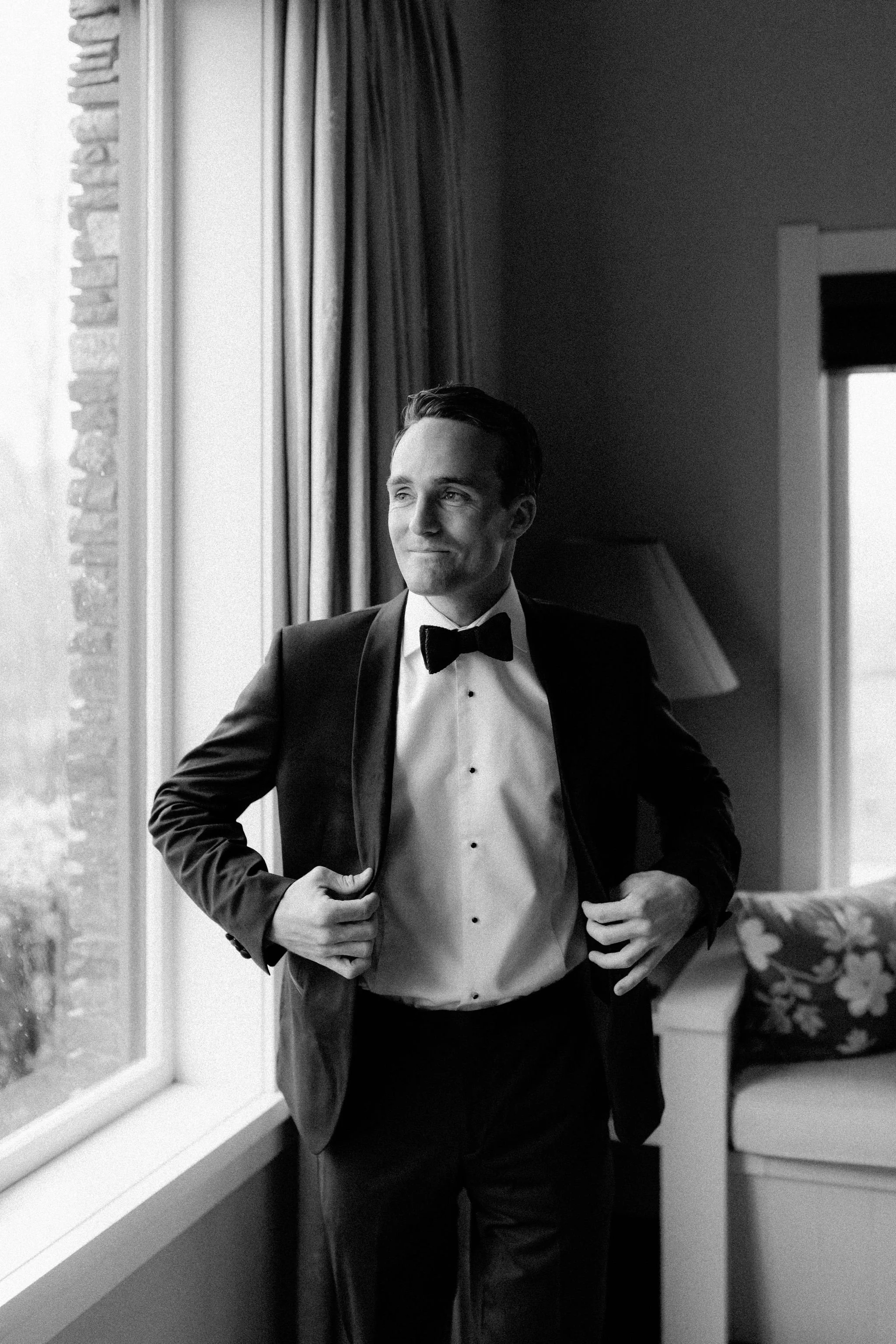 A man dressed in a tuxedo with a bow tie is standing indoors near a window, looking out thoughtfully.