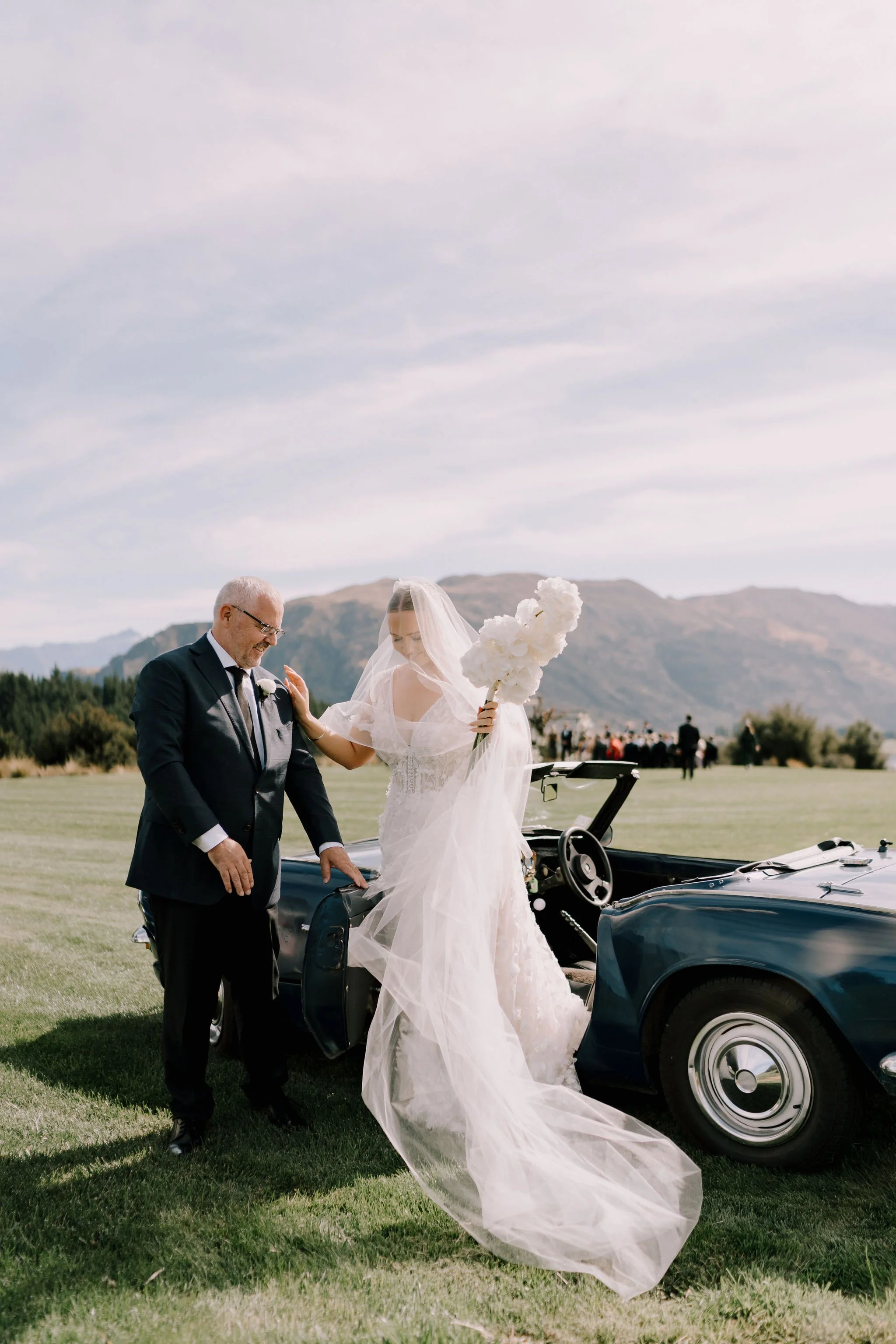 A bride in a wedding dress and veil holding a bouquet exits a vintage car with an older man, likely her father, on a grassy field with a mountain landscape in the background.