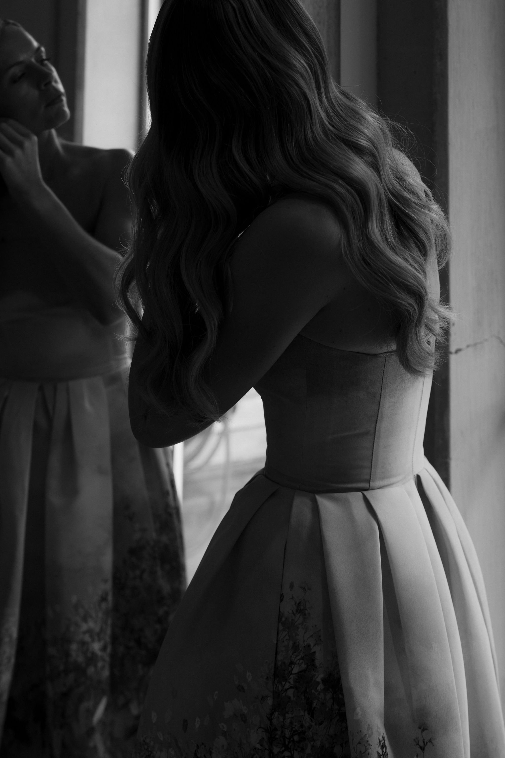 A woman in a strapless dress adjusting her hair in front of a mirror, with another woman helping her in the background.