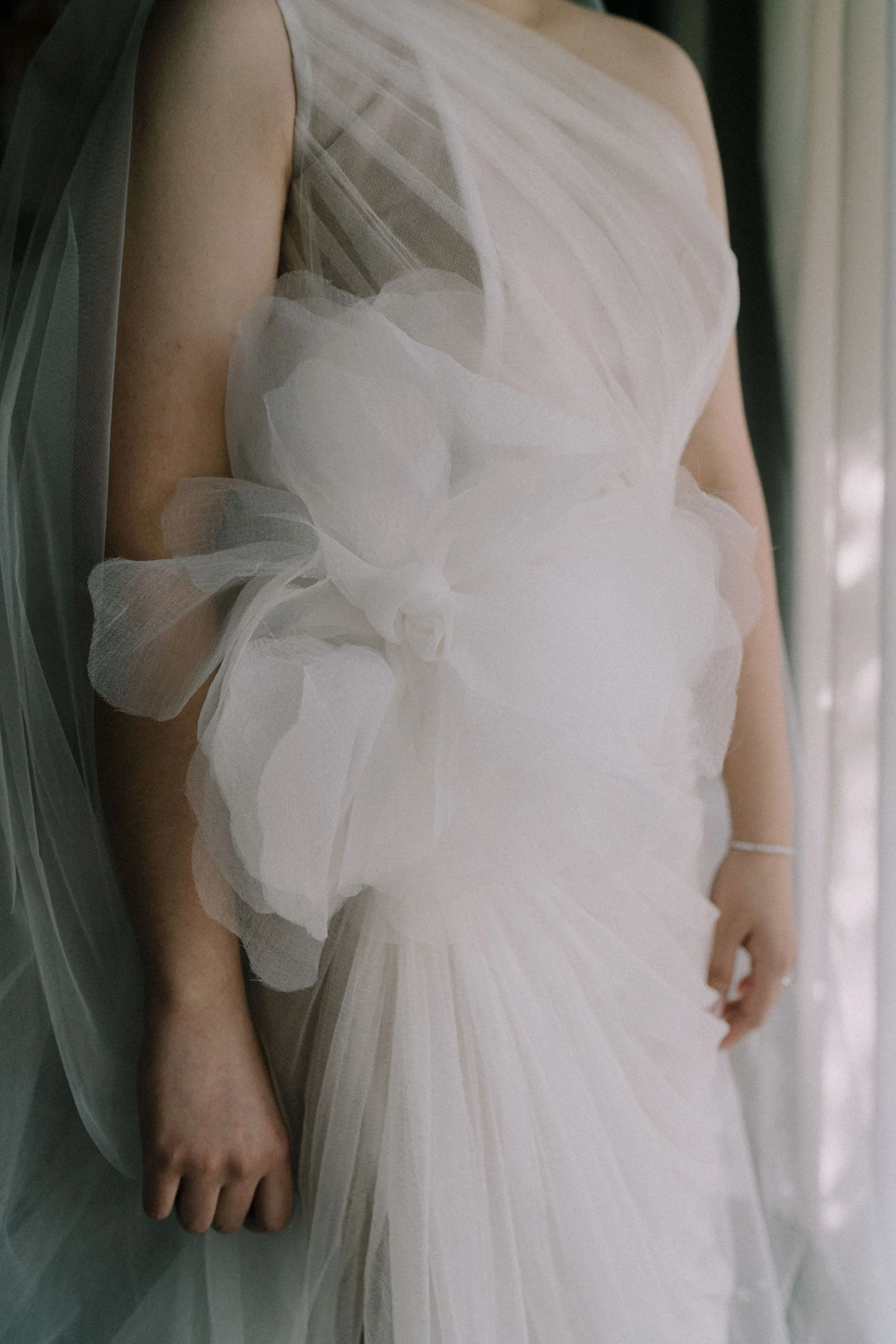 Close-up of a person wearing an elegant white wedding dress with sheer, layered fabric and a large bow detail on the waist, standing near a window.