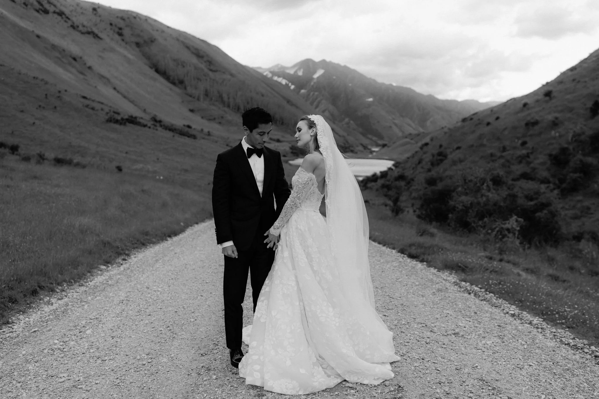A bride and groom standing on a gravel path in a mountainous landscape, holding hands and looking at each other, black-and-white photo.