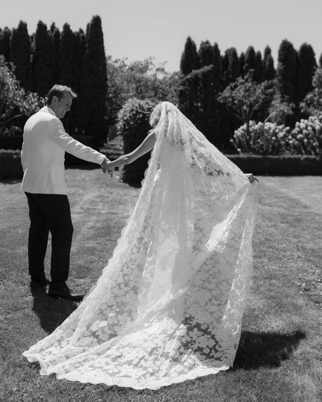 A bride and groom holding hands and smiling in an outdoor garden, with the bride wearing a lace wedding dress and veil, and the groom in a white tuxedo jacket, surrounded by trees and shrubbery.