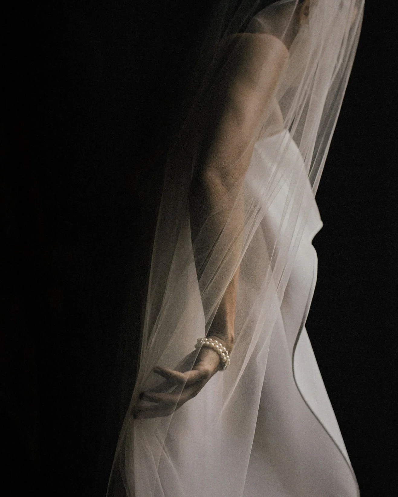Close-up of a woman's hand wearing pearl bracelets, with sheer fabric and a white dress, against a dark background.