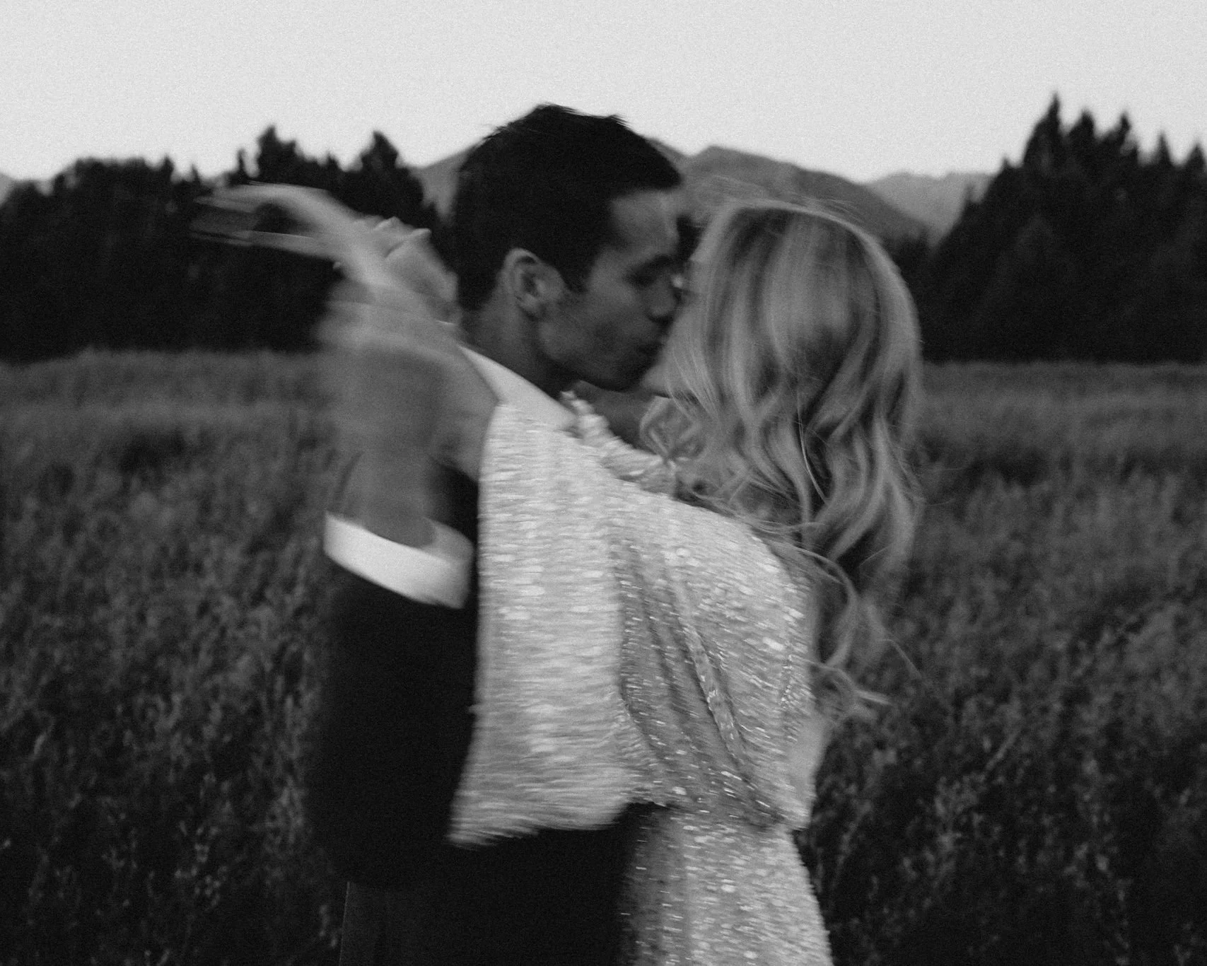 A black and white photo of a couple kissing outdoors in a field with mountains in the background.