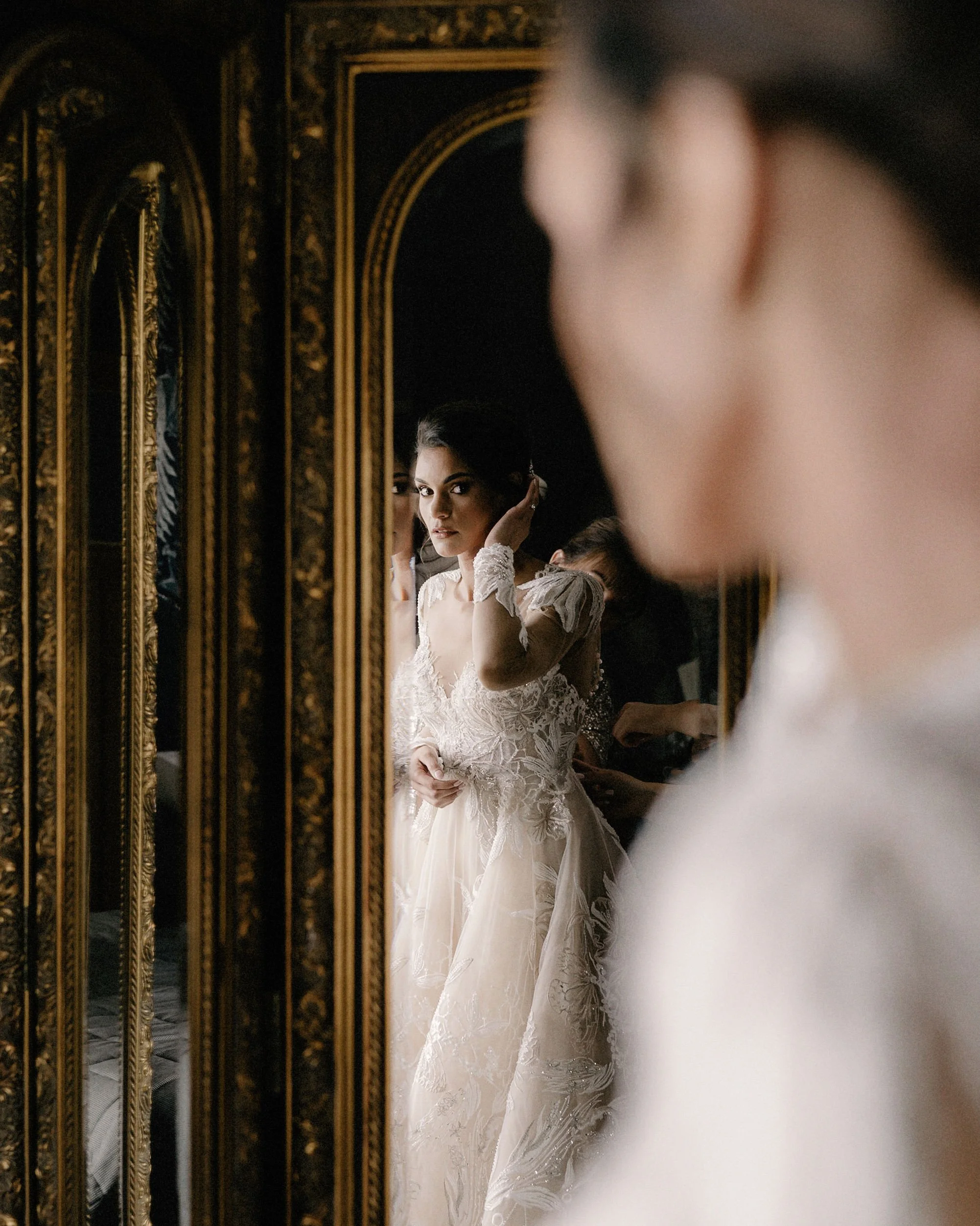A bride looking at herself in a mirror, adjusting her hair, wearing a lace wedding dress with intricate embroidery, in a room with ornate gold-framed mirrors.