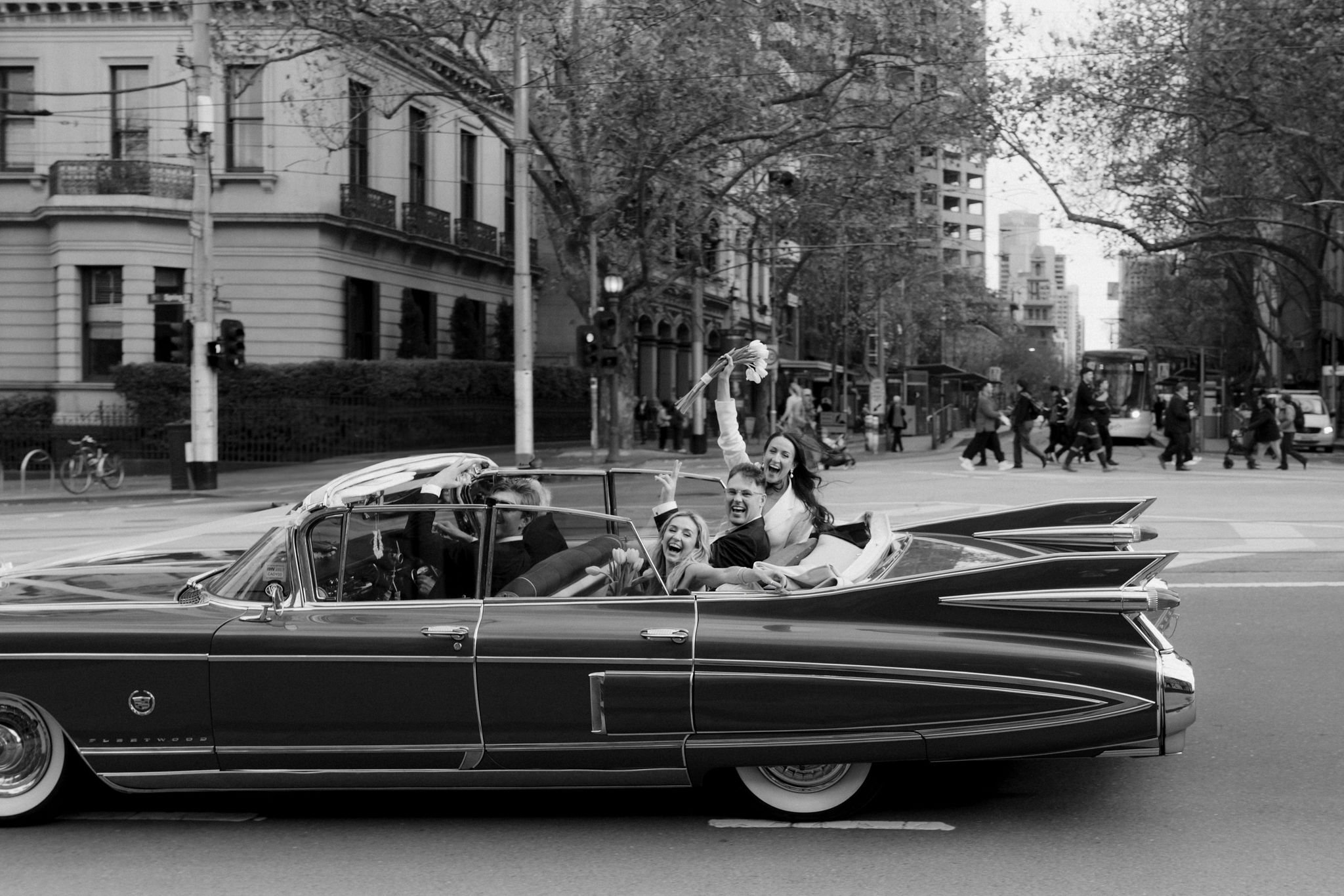 A group of people celebrating on a vintage Cadillac car in an urban street with buildings, trees, and pedestrians in the background.