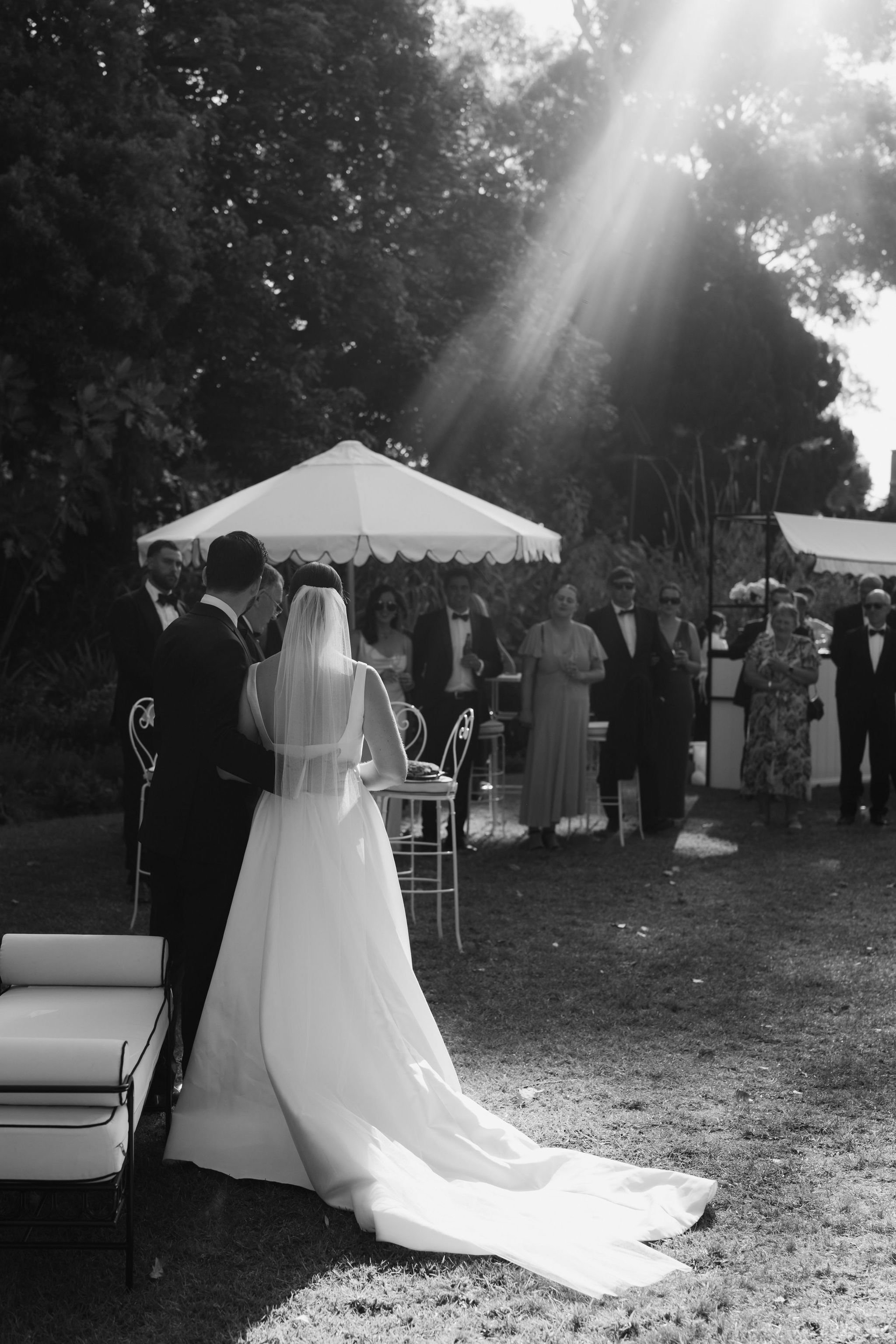 Black and white photo of a wedding ceremony outdoors under trees, with the bride and groom at the altar. The bride wears a white gown with a long train and veil; the groom wears a dark suit. Guests stand nearby, some wearing sunglasses, against a backdrop of trees and a white umbrella.