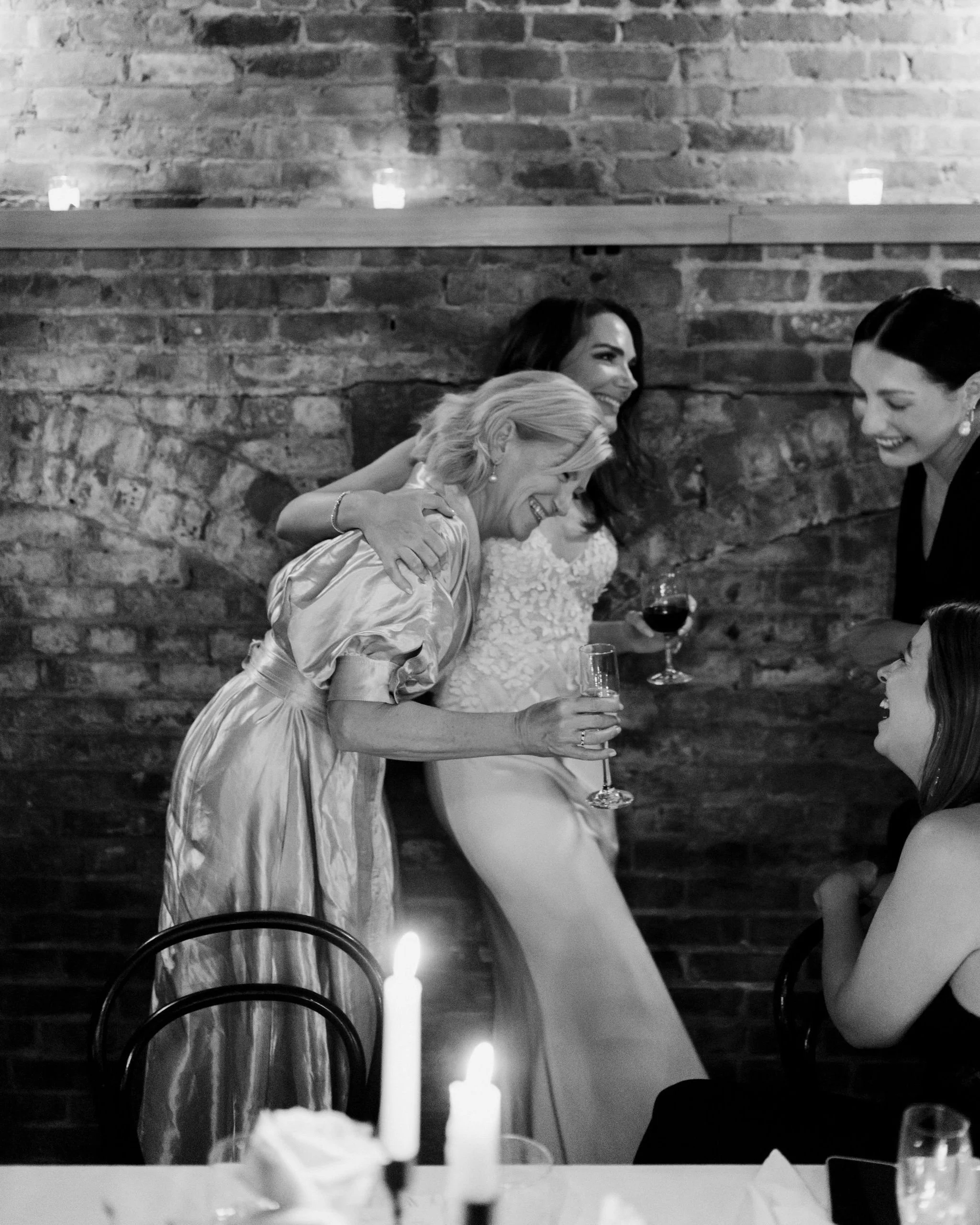 A group of women at a celebration, smiling and laughing, with two women standing and holding glasses of wine, one of whom is embracing another woman who is sitting, in a room with a brick wall and candles.